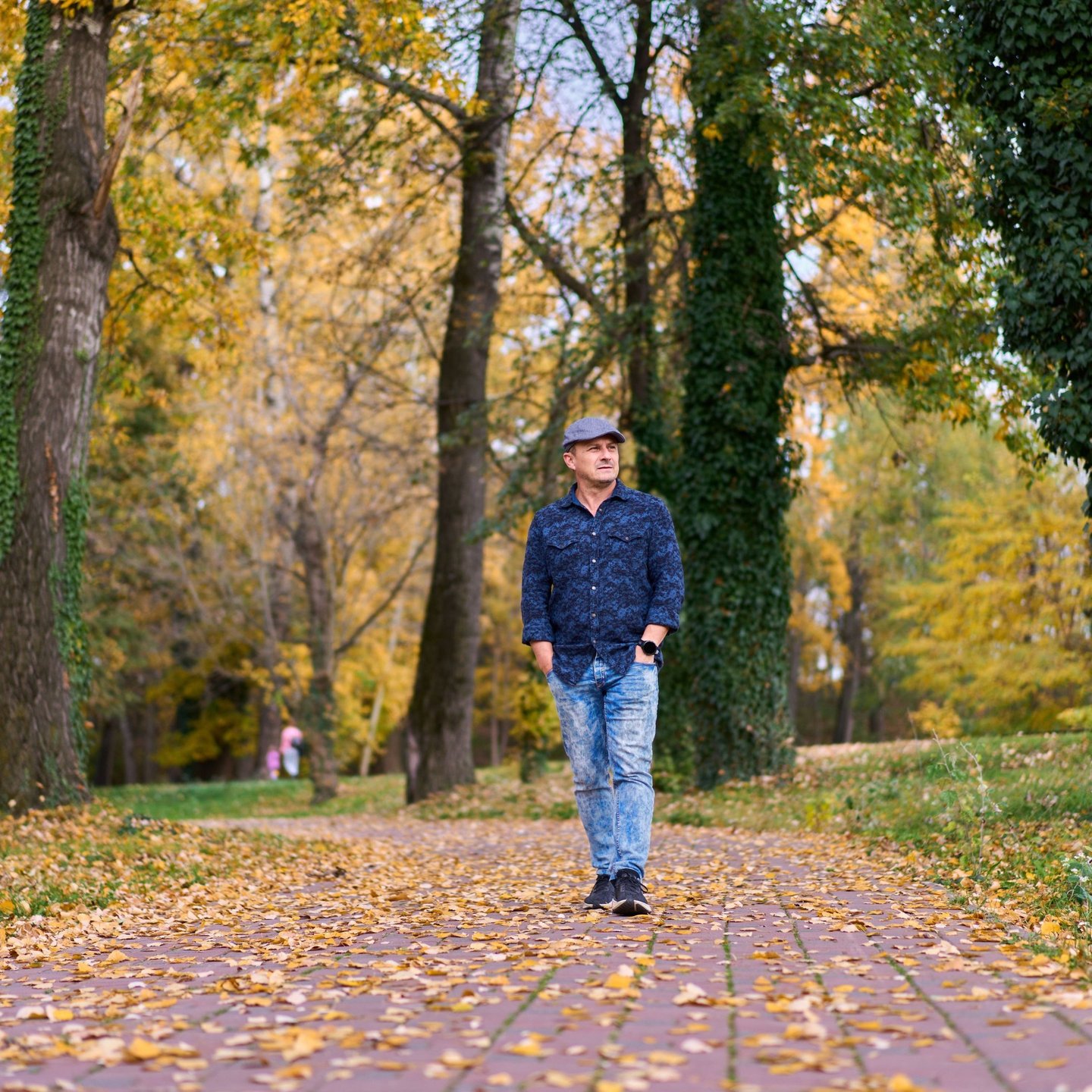 A man walking along a paved path in a Calgary park on a fall afternoon, hands in jacket pockets, contemplative expression