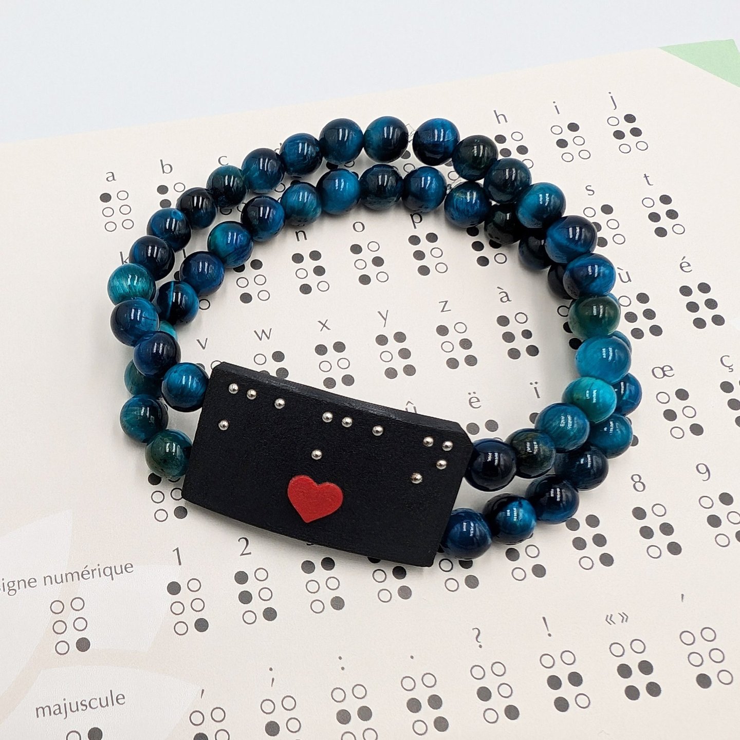 Bracelet perle bleue avec une plaque en braille et un cœur rouge, posé sur un alphabet braille.