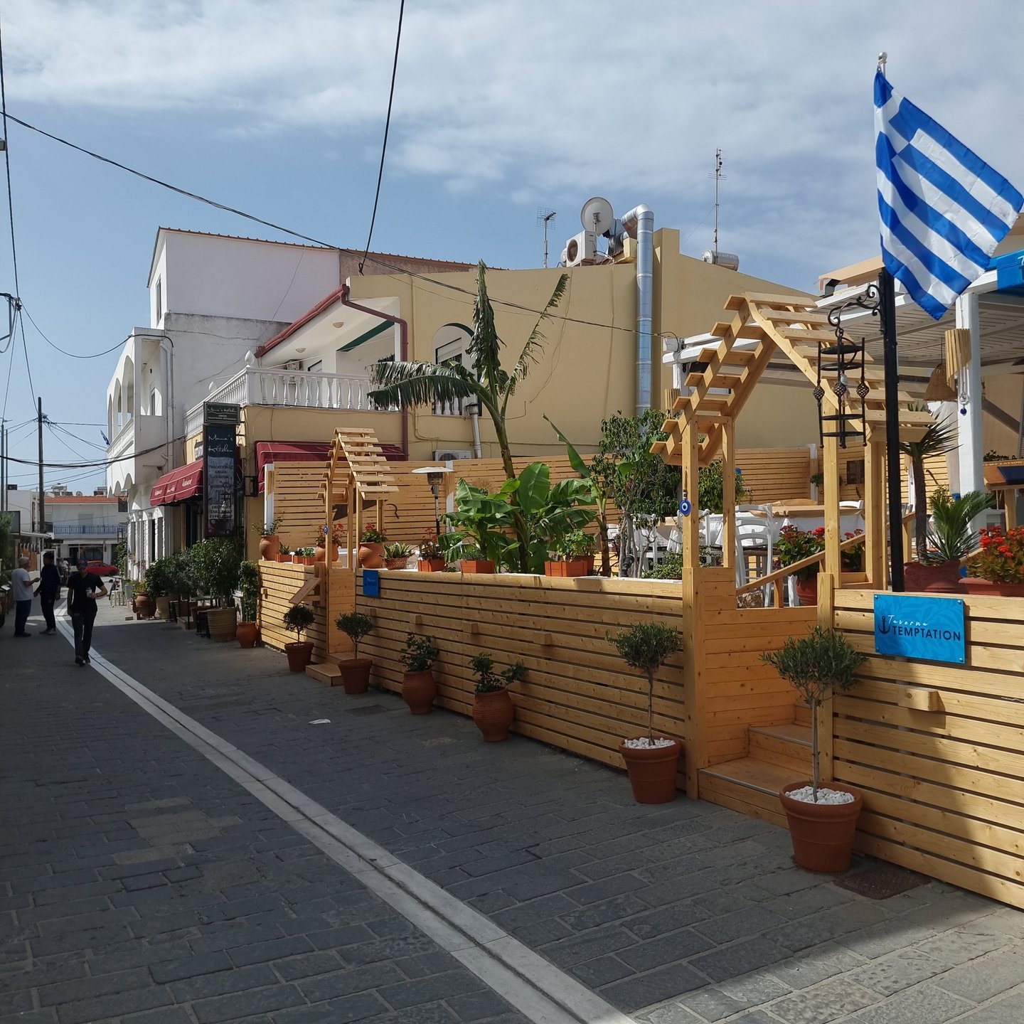 Greek tavern outdoor seating area with wooden fencing, potted plants, and a Greek flag on a cobblestone street.
