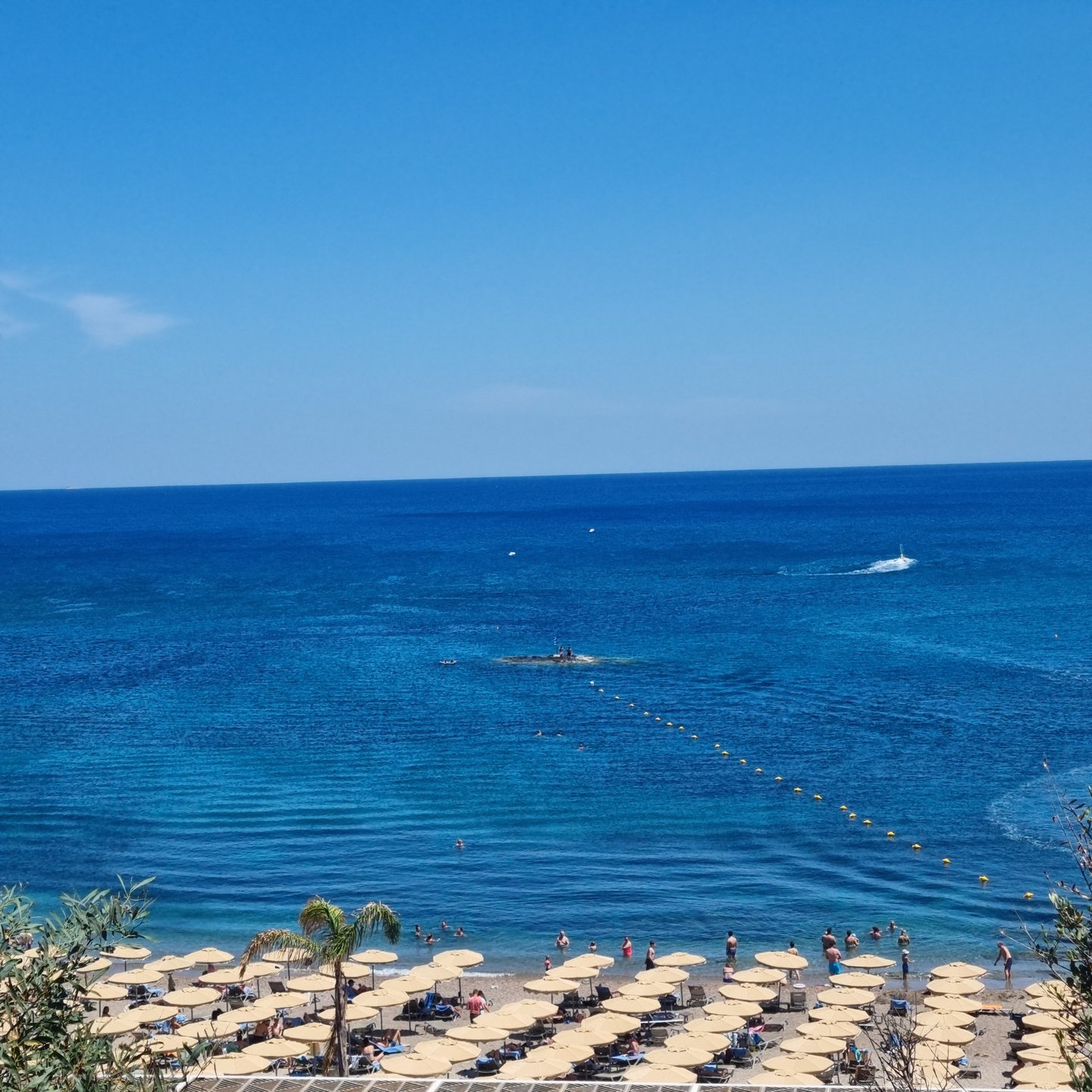 Wide view of a sunny beach resort with tan umbrellas and people swimming in deep blue ocean water.