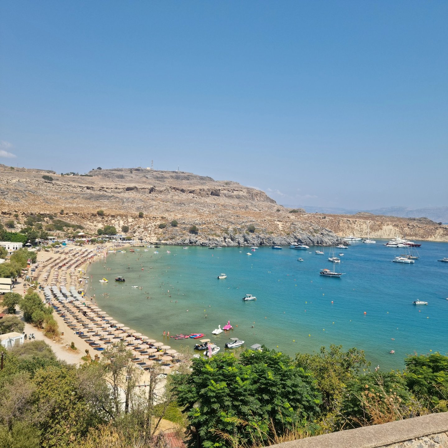 Panoramic view of Lindos beach in Rhodes with turquoise water, rows of umbrellas, and boats at anchor.