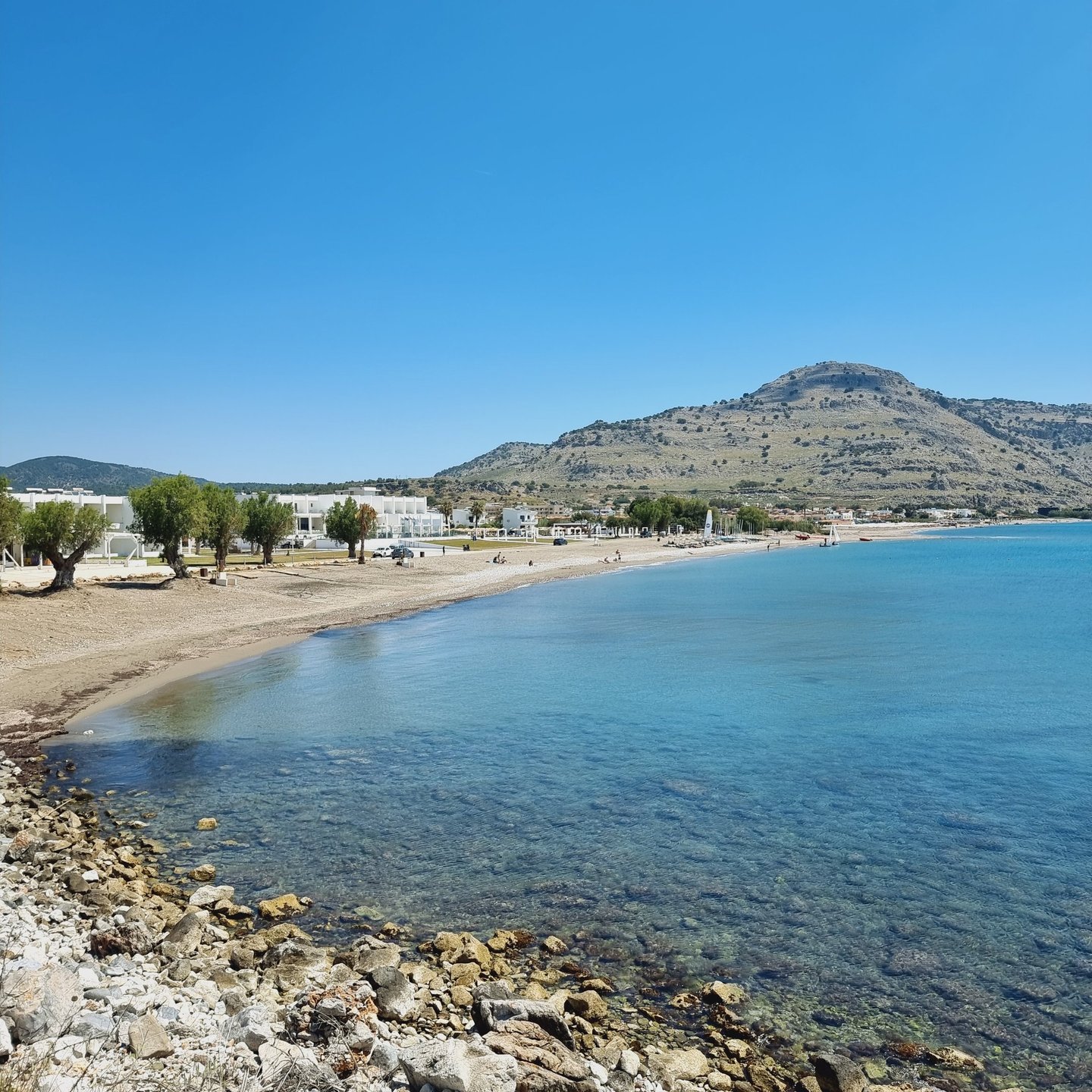 Panoramic view of a sandy Greek beach with turquoise sea, white buildings, and coastal mountains under a clear blue sky.