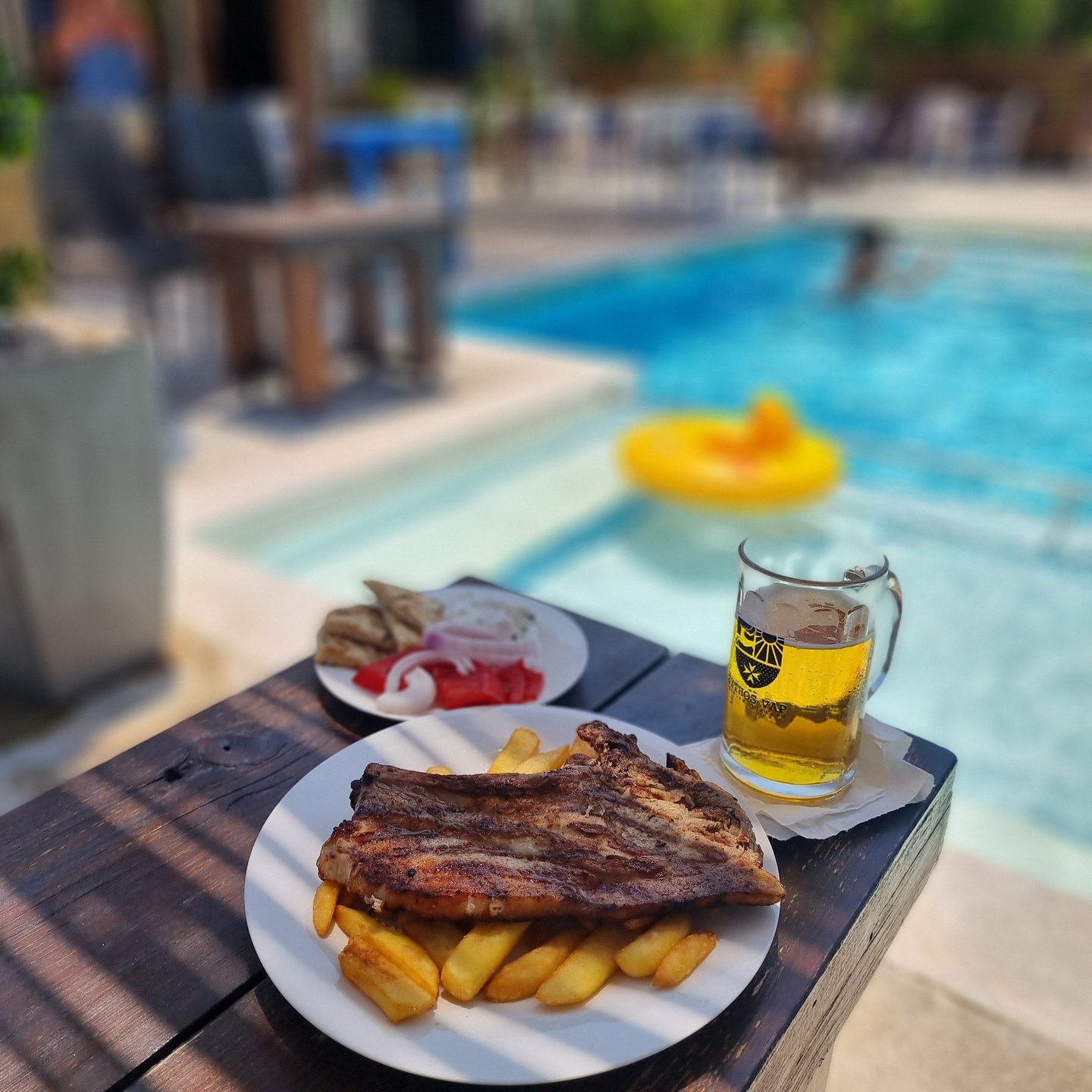 Grilled pork steak and french fries served with cold beer by a sunny swimming pool.