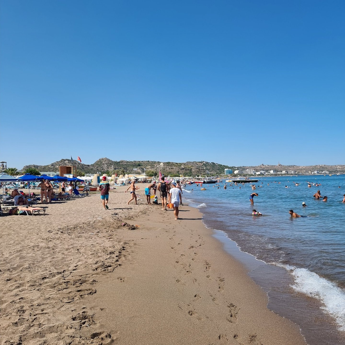 Crowded sandy beach in Rhodes, Greece, with tourists swimming in the blue Mediterranean Sea under a clear sky.