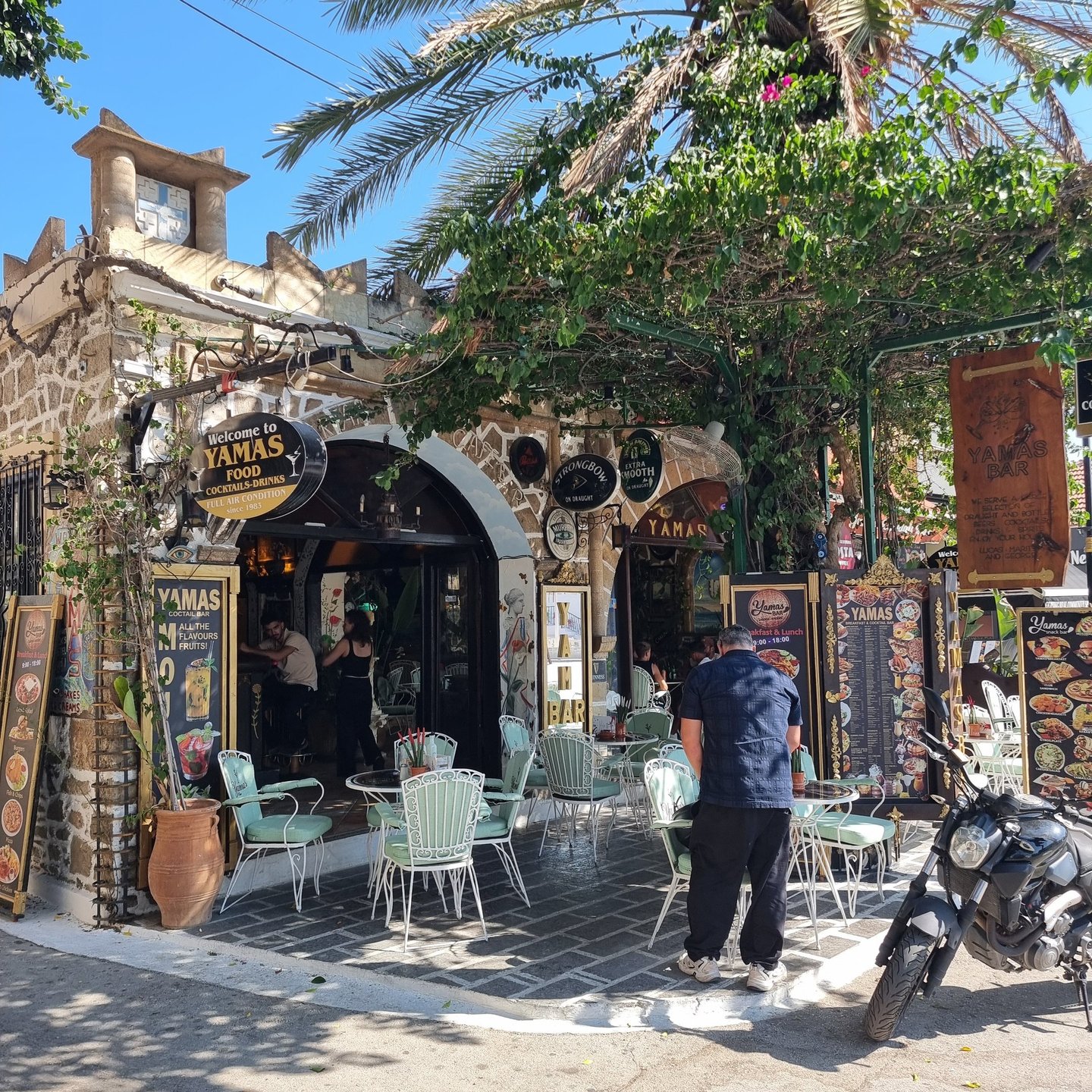 Charming outdoor seating area at Yamas bar and restaurant with Greek stone architecture and palm trees.