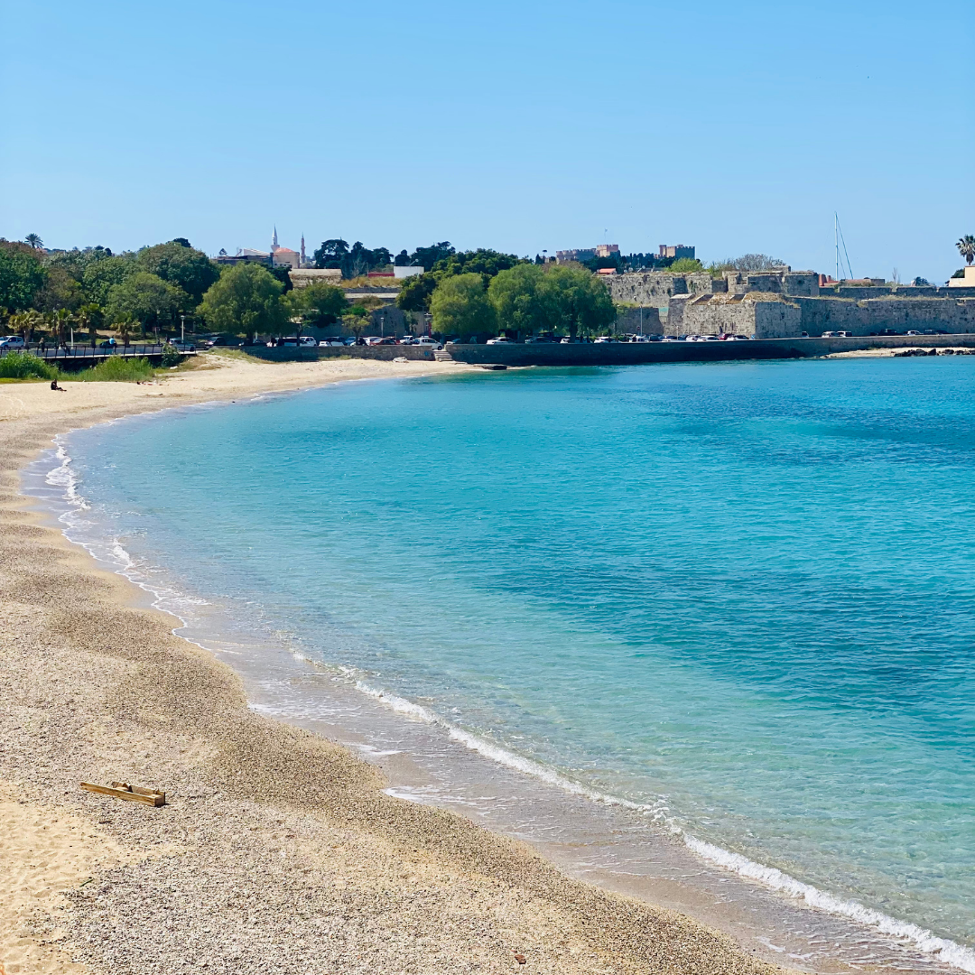 Sunny Rhodes beach with turquoise water, sand, and historic stone walls in the background under a blue sky.