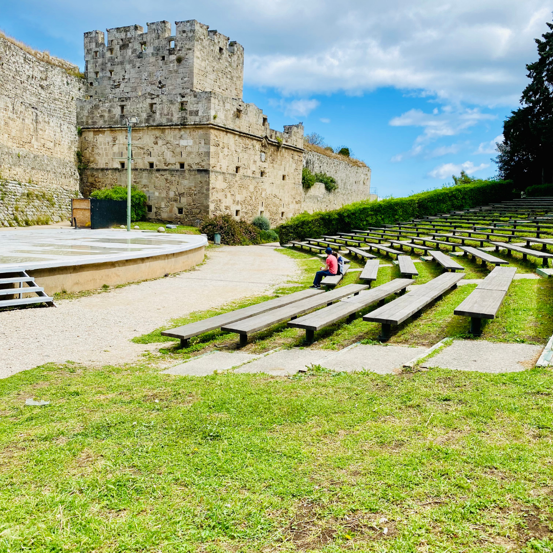 Outdoor stone amphitheater and historic castle walls in Rhodes, Greece, under a blue sky.