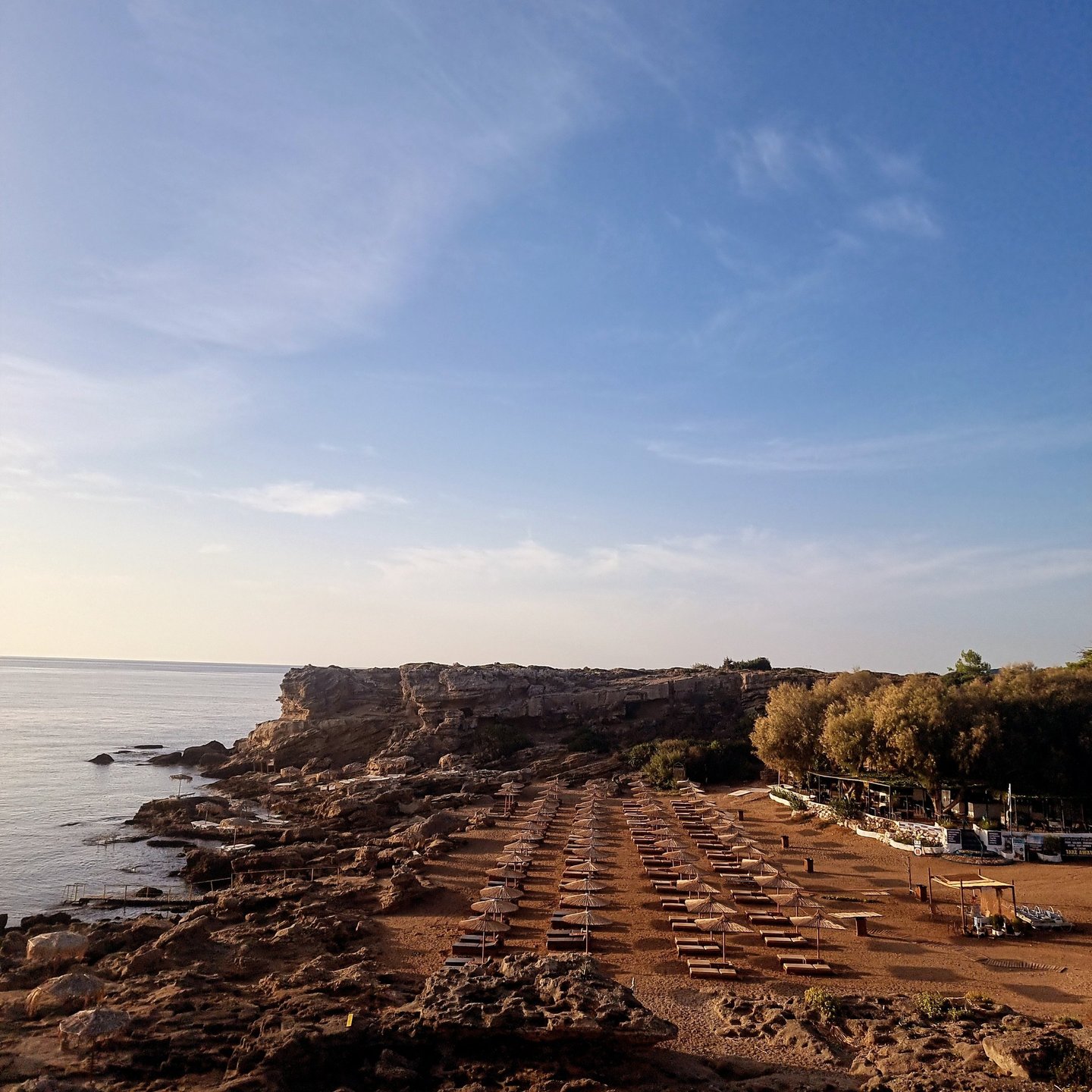Seaside beach club with rows of sun loungers and umbrellas on a rocky coastline under a clear blue sky.