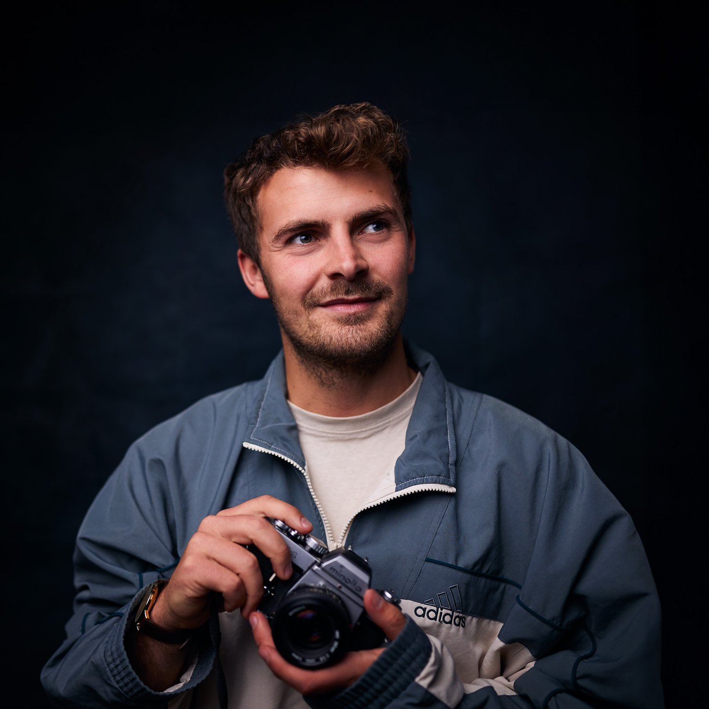 A smiling man holding a vintage Minolta film camera while wearing an Adidas jacket.