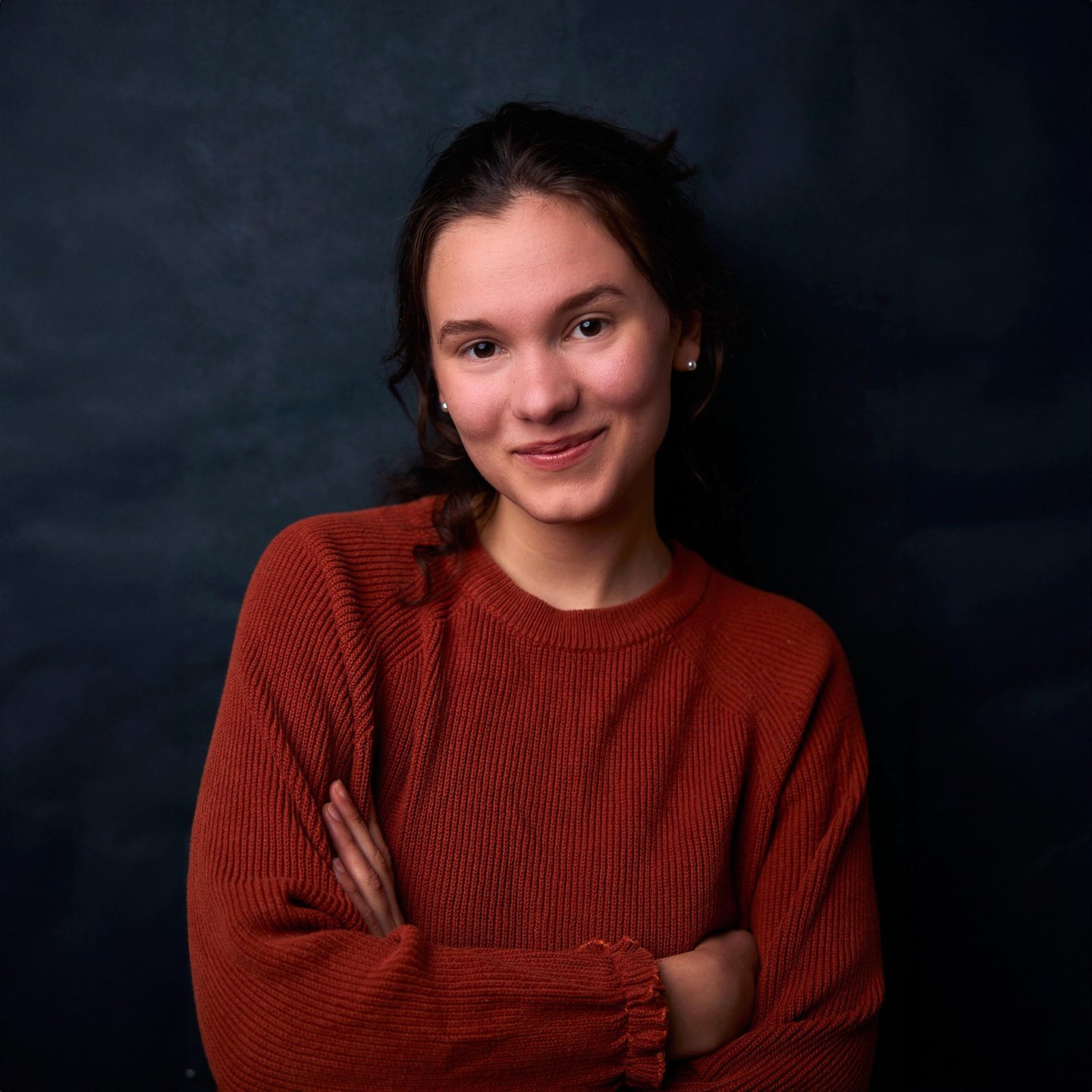 A smiling young woman posing in a rust-colored knit sweater against a dark backdrop.