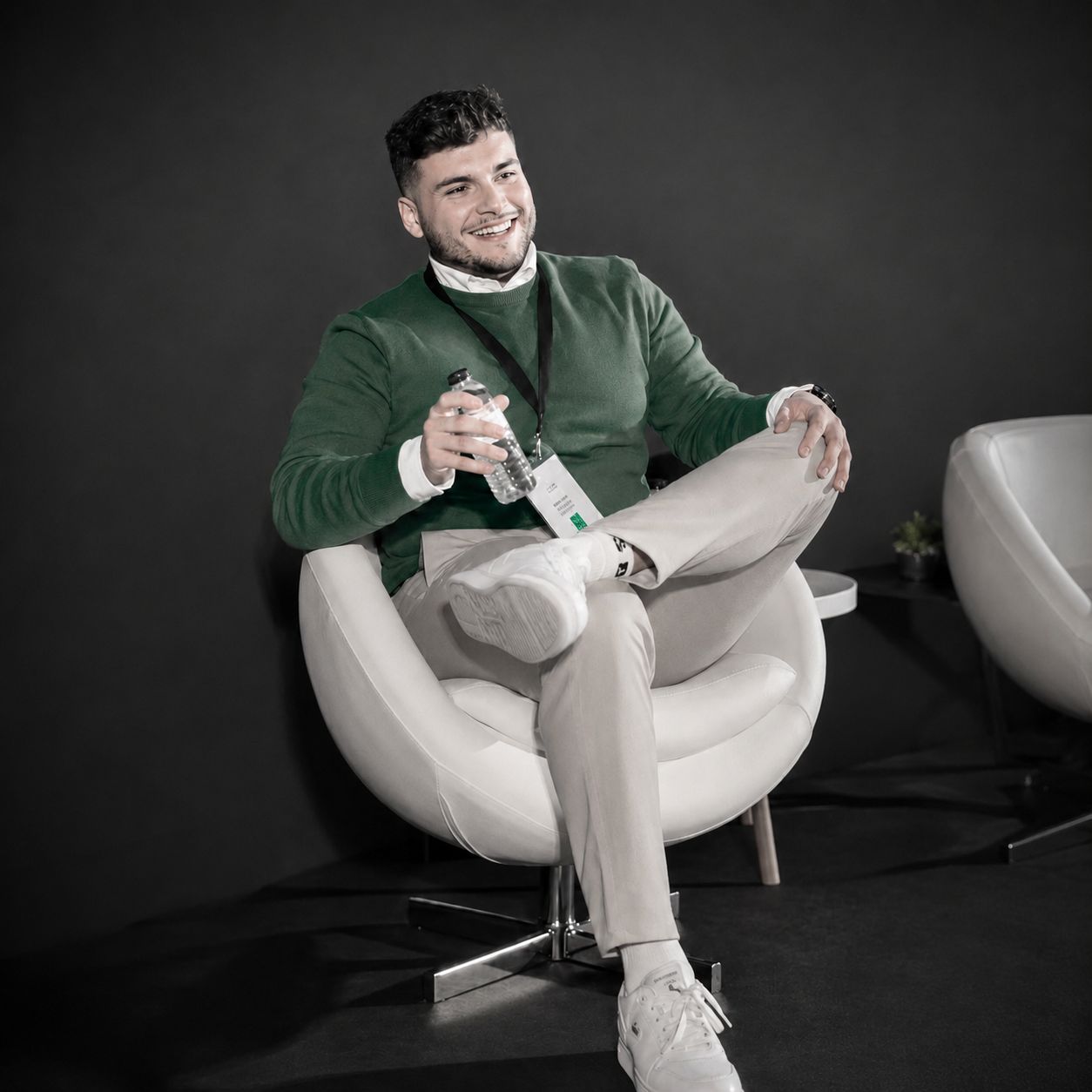 Smiling man in a green sweater sitting on a white swivel chair at a professional event conference.