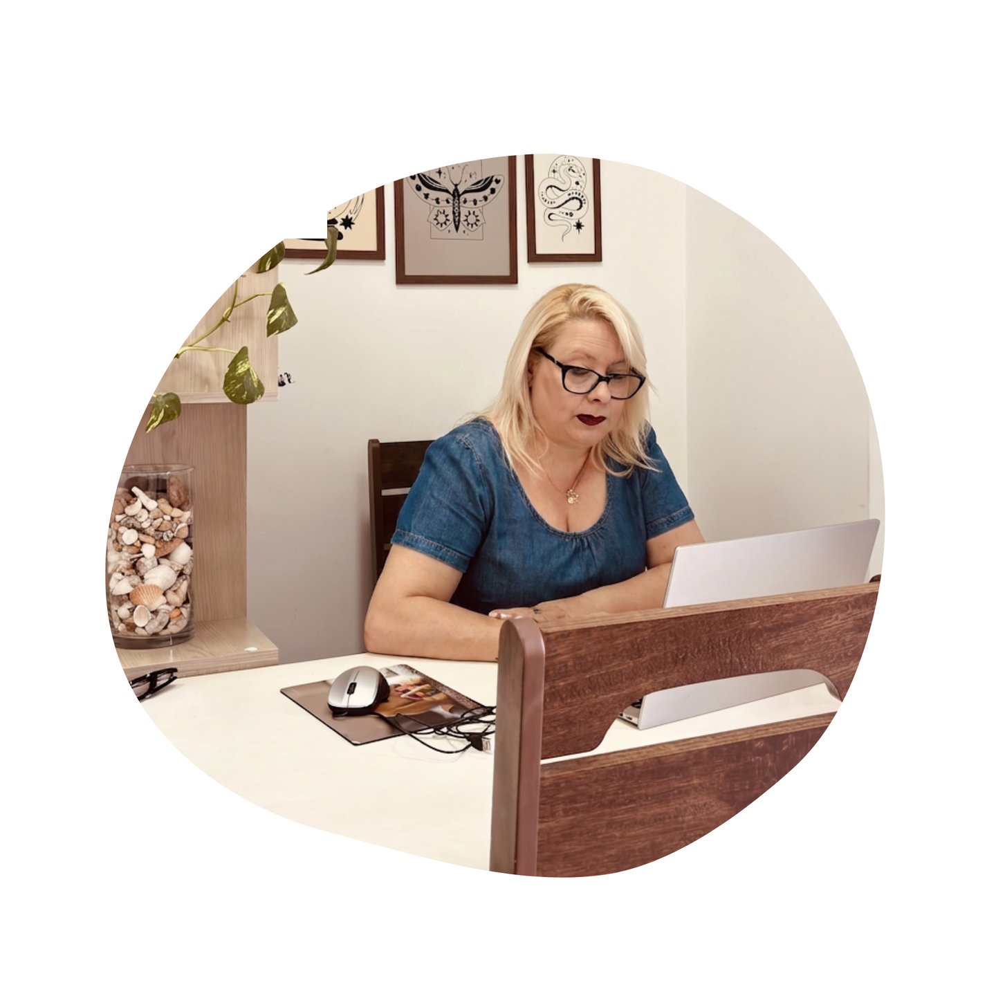 A blonde woman wearing glasses works on her laptop at a home office desk with modern decor.