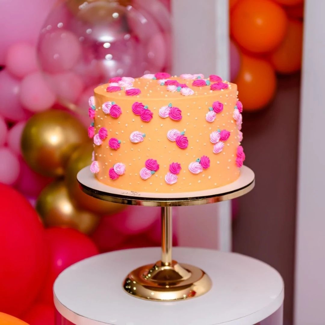 Bolo decorado com rosas coloridas em um suporte dourado, rodeado por balões em tons vibrantes. Ideal