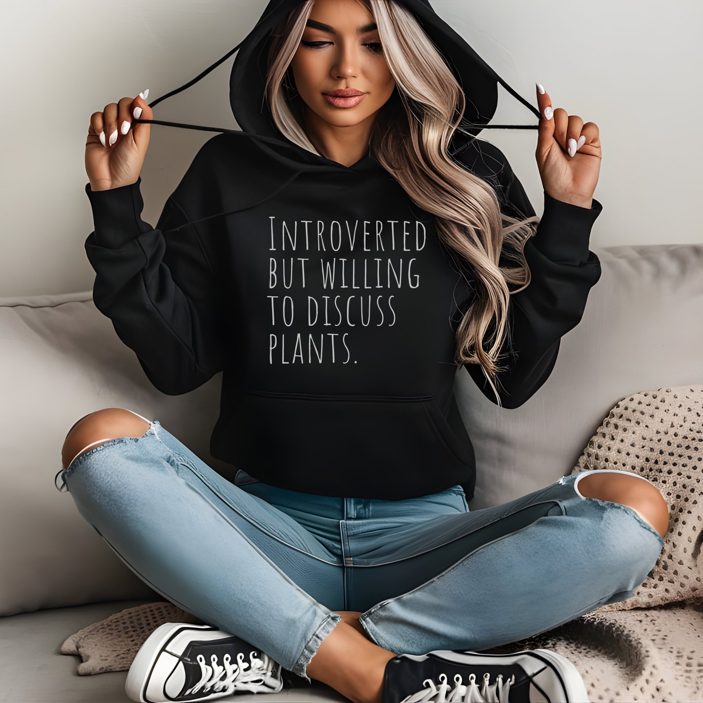 Woman in a black “Introverted but willing to discuss plants” hoodie sitting cross-legged indoors.