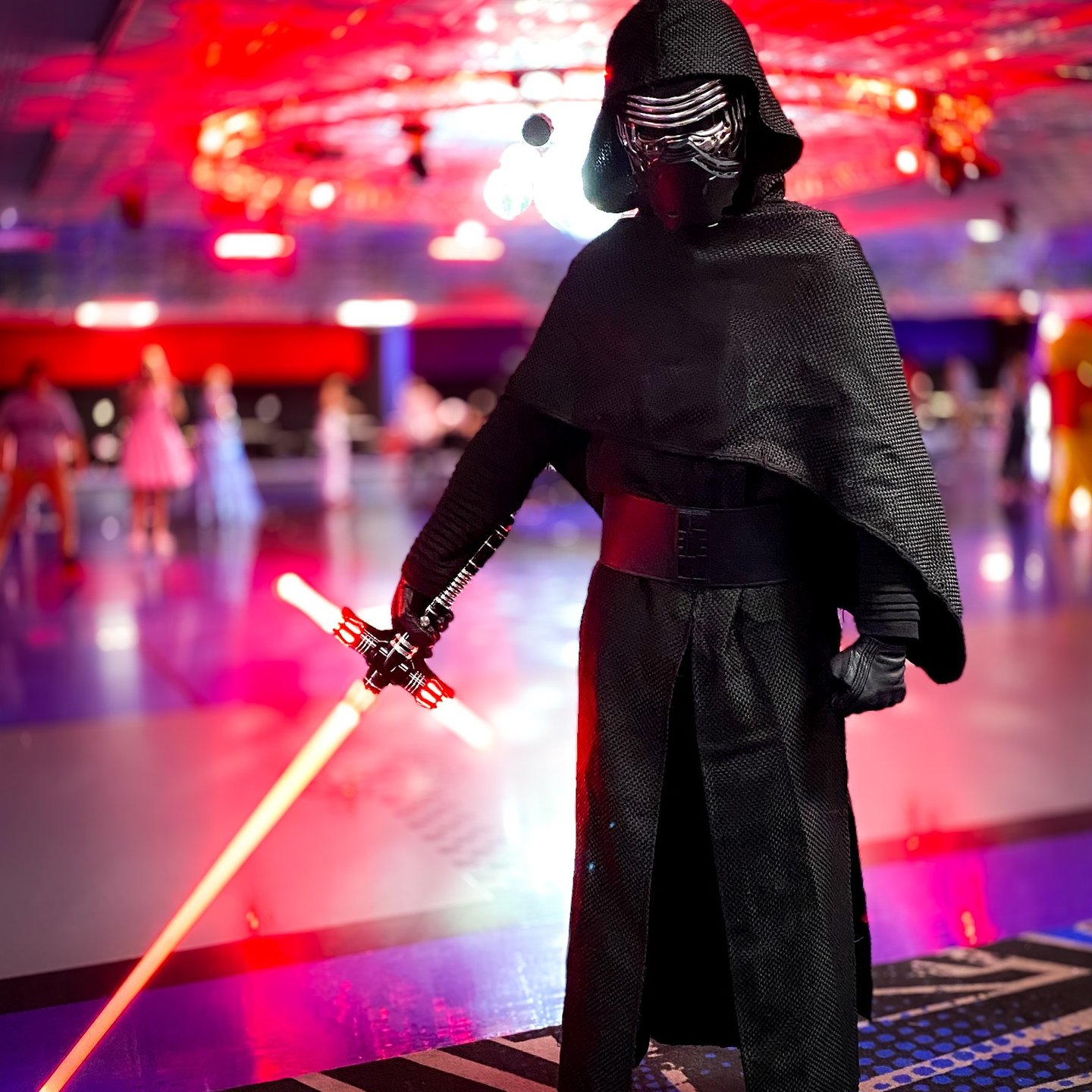 A person in a Kylo Ren cosplay costume holding a glowing red crossguard lightsaber at a roller rink.
