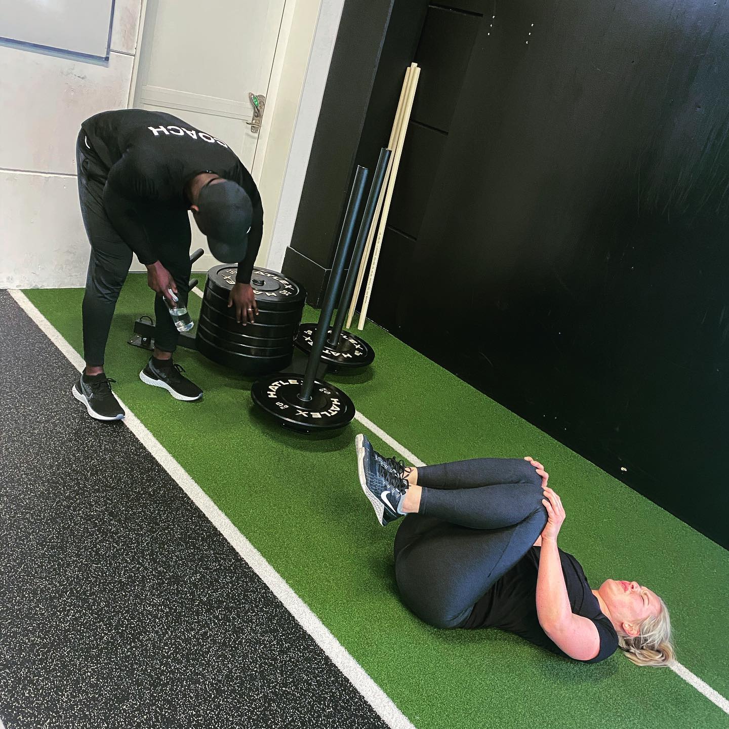Personal trainer assisting a woman performing knee-to-chest stretches on gym turf during a workout.