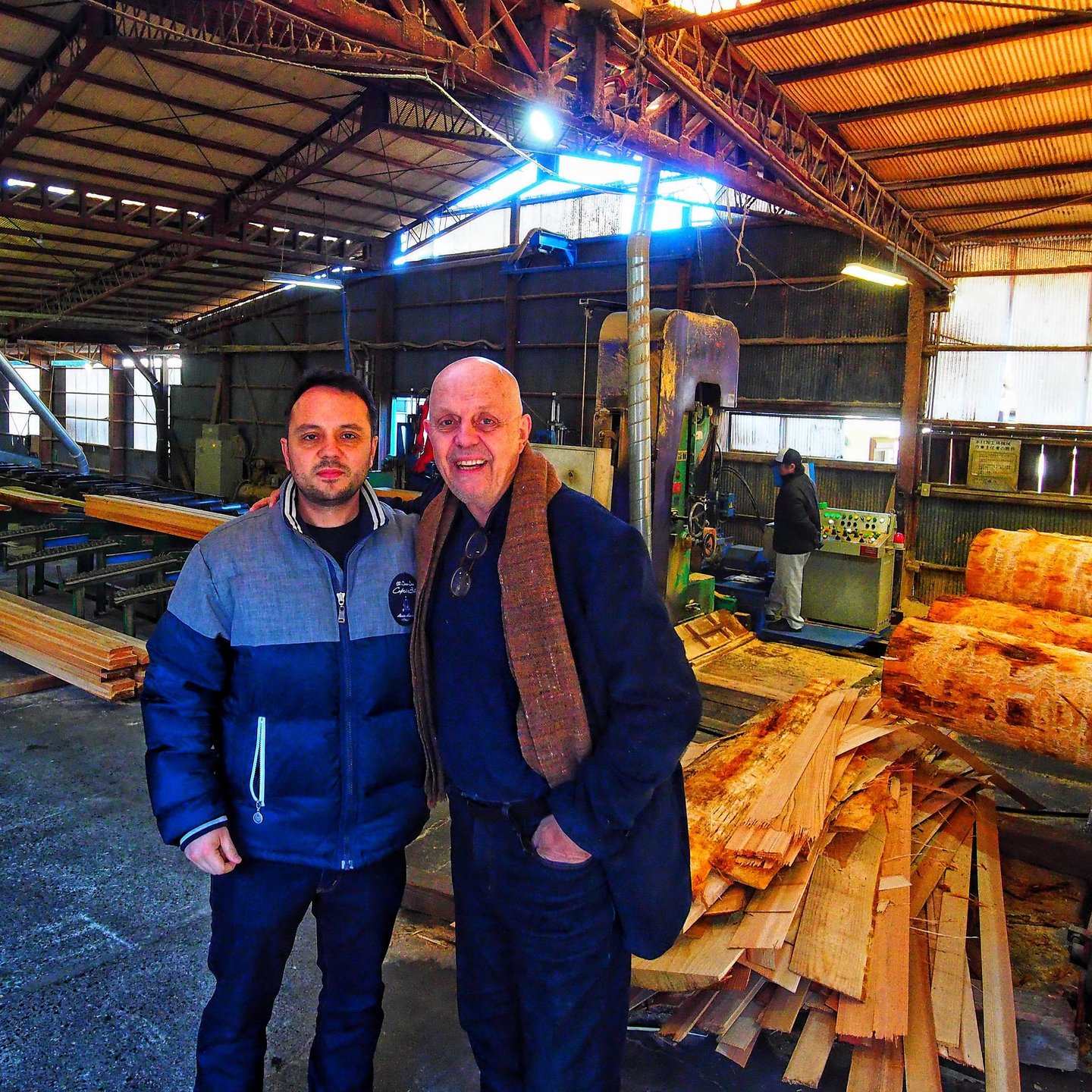 Two men standing inside a woodworking factory or sawmill surrounded by stacks of cut lumber and logs.