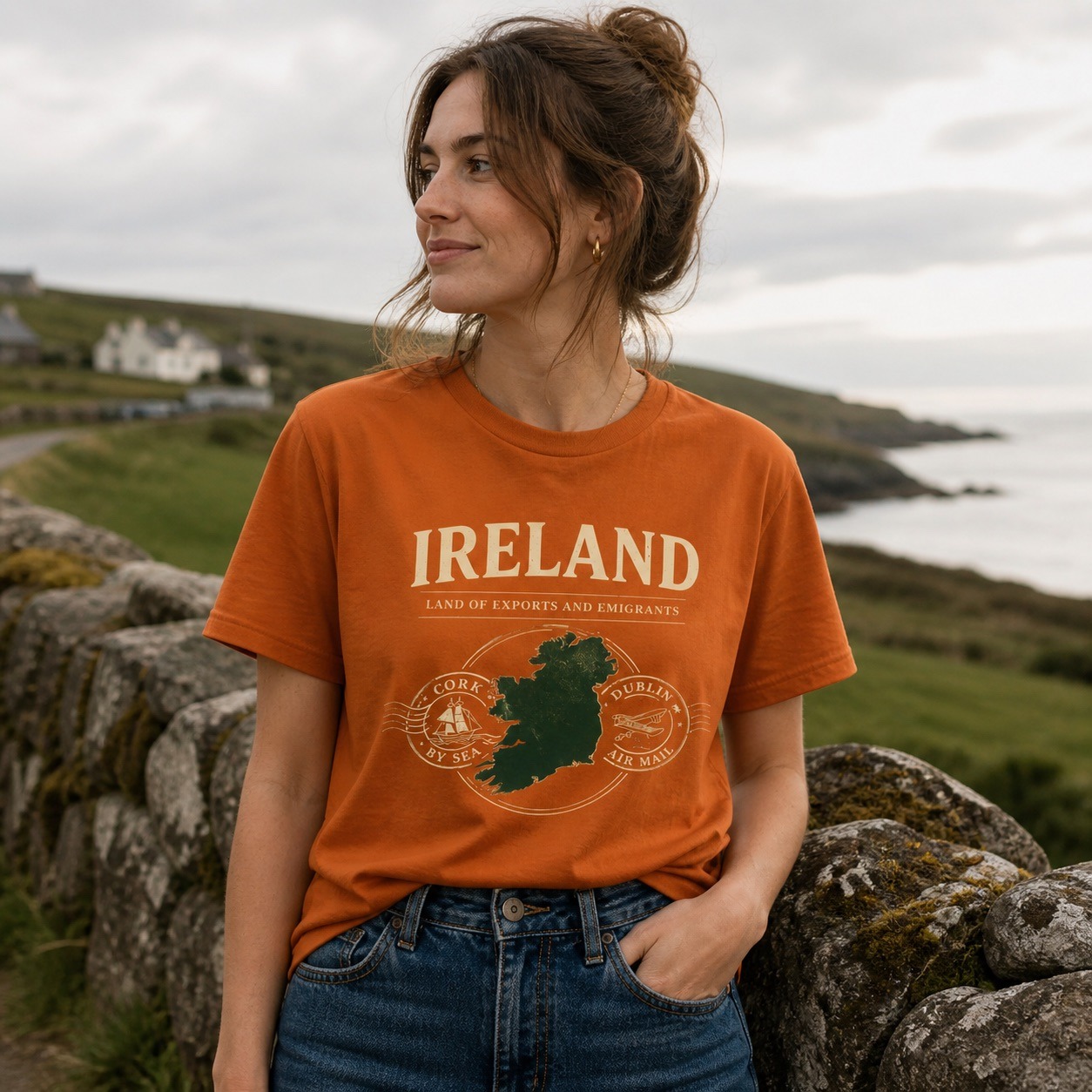 Woman wearing an orange Ireland graphic t-shirt by a coastal stone wall.