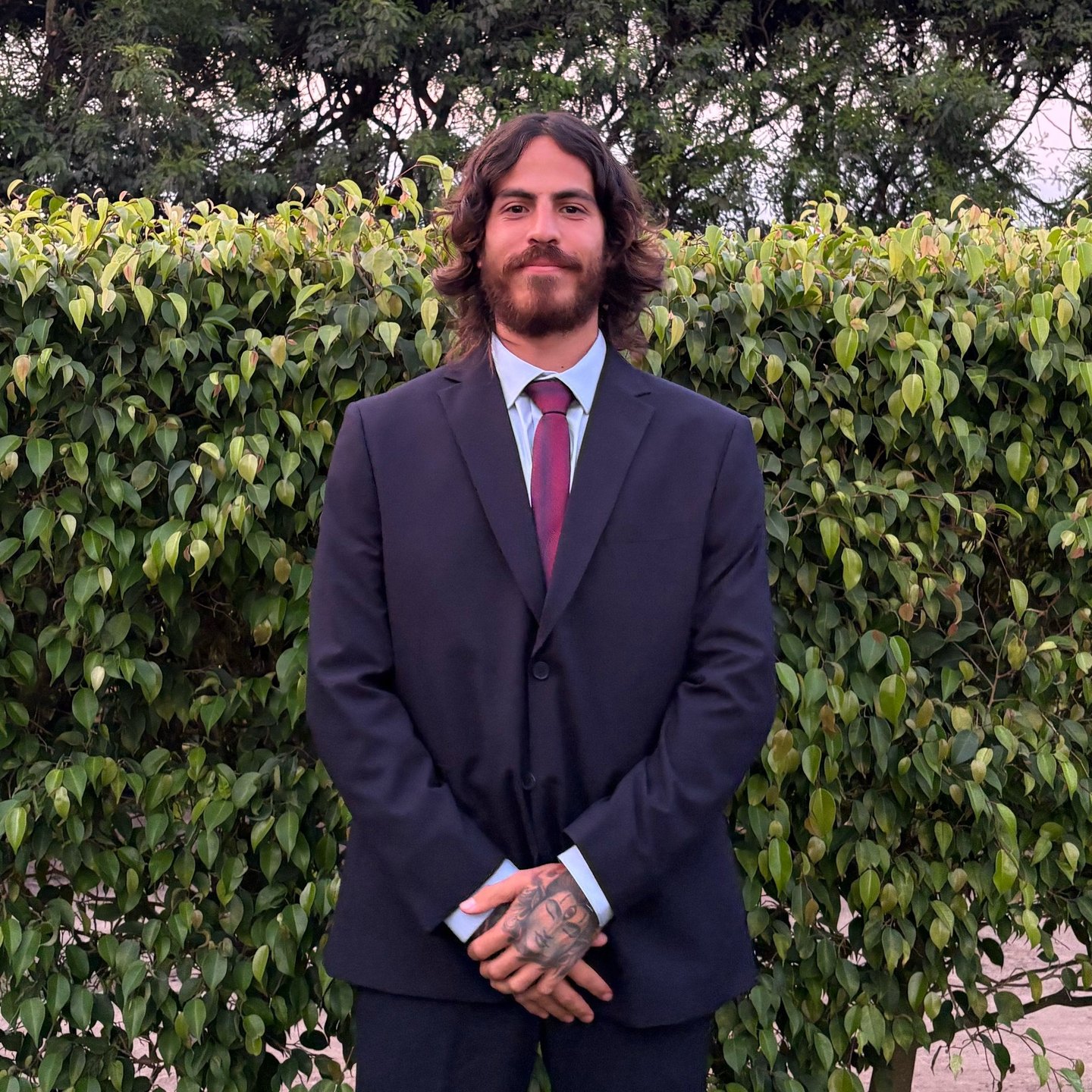 A smiling man with long wavy hair and a beard wearing a formal suit and tie outdoors.