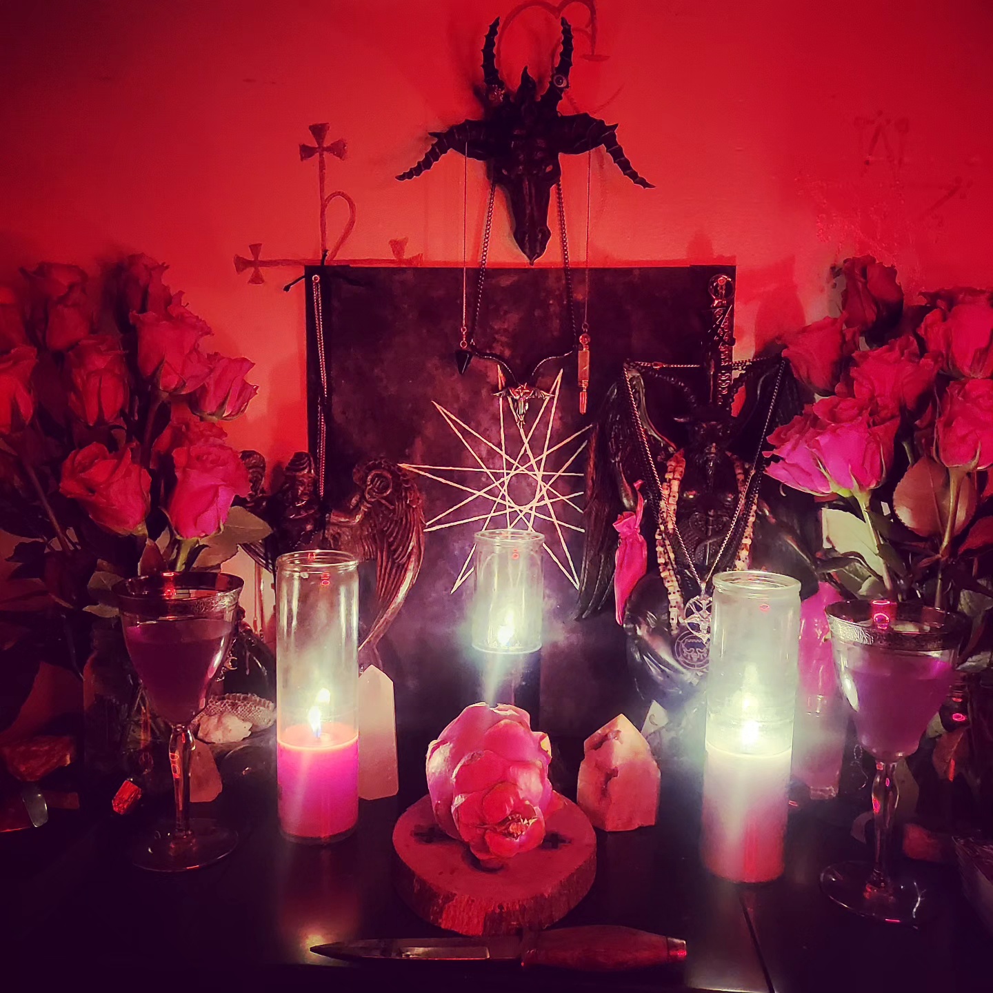 Satanic altar with Baphomet sigil, red roses, lit ritual candles, and occult symbolism under red light.