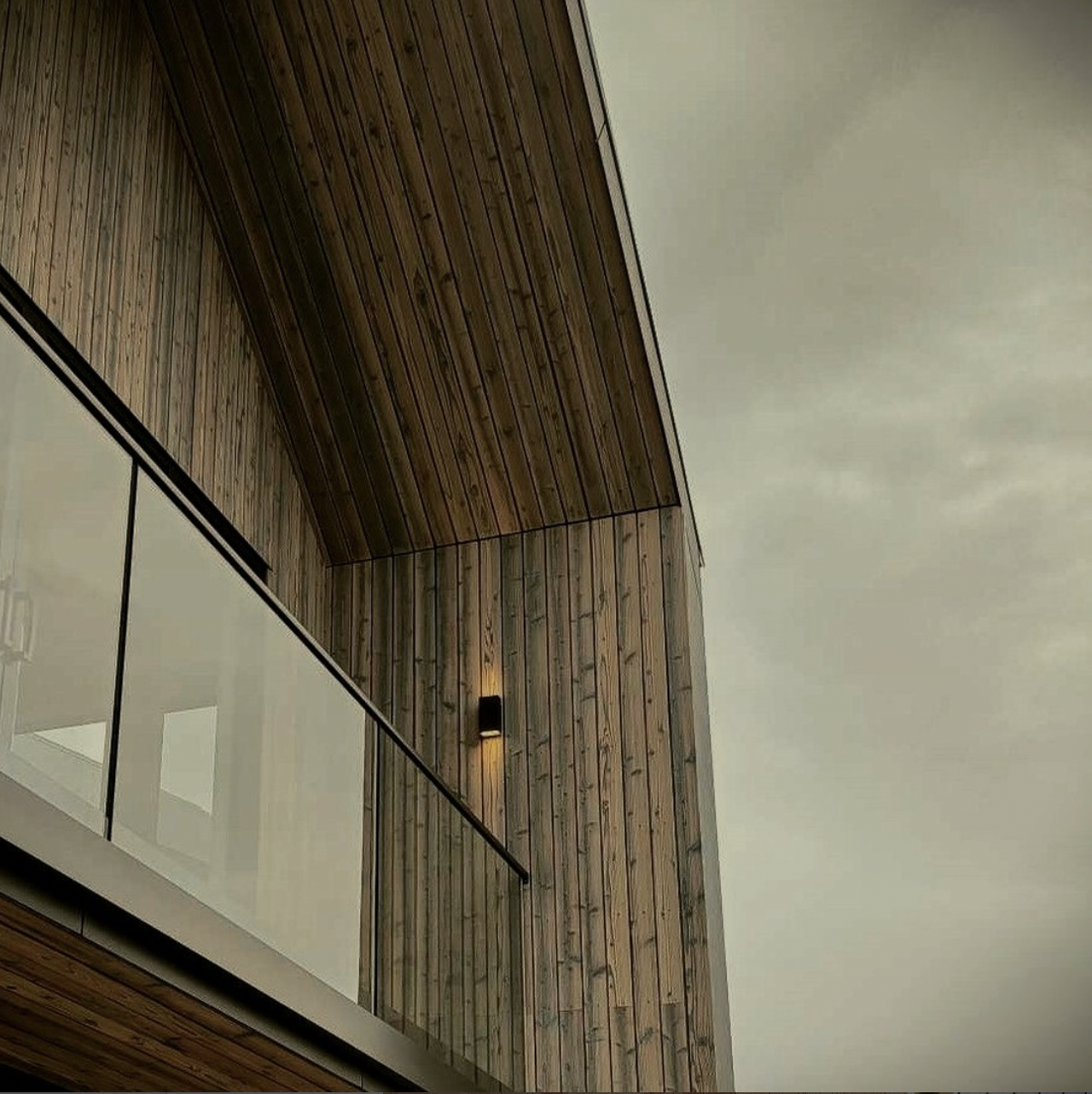Modern balcony featuring vertical cedar wood siding, glass railing, and exterior wall lighting under a moody sky.