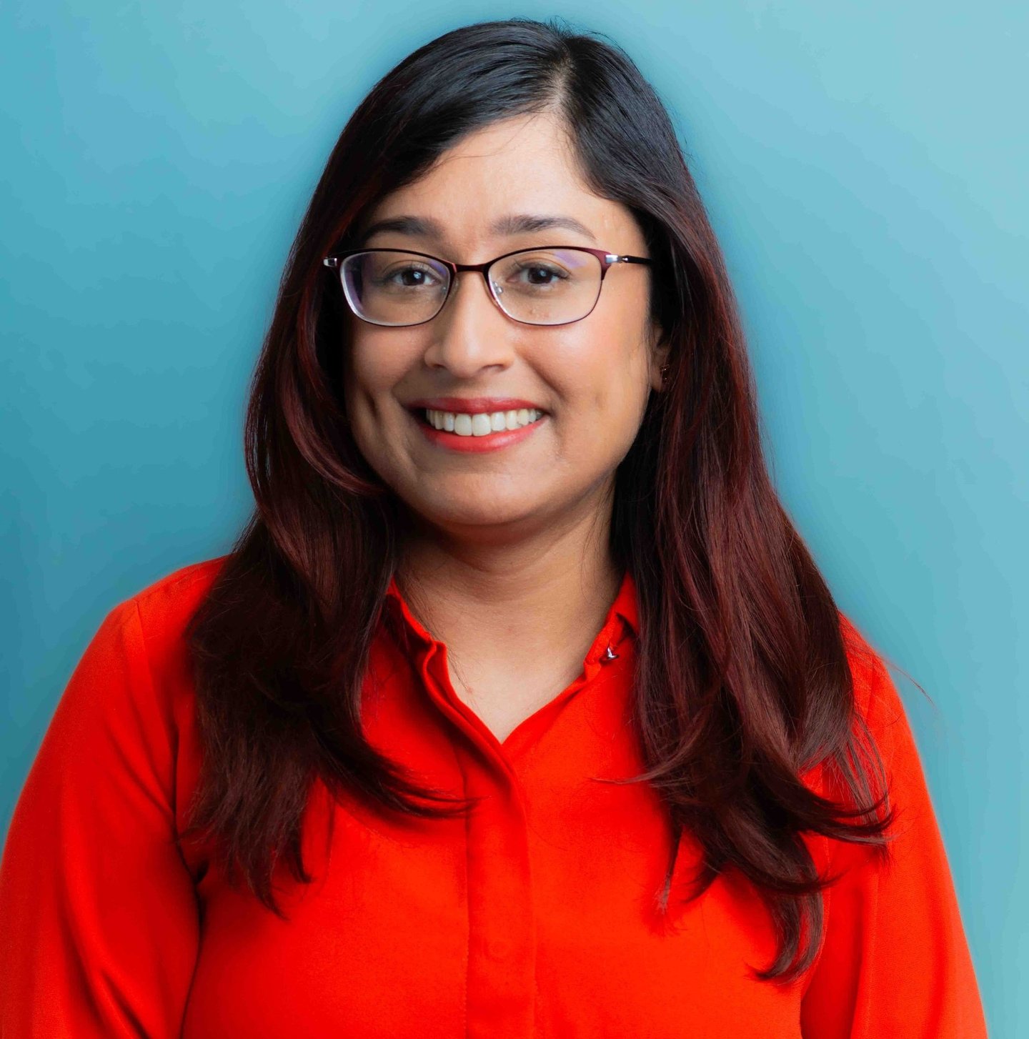 A smiling woman in glasses stands against a blue background, wearing a vibrant red shirt and a friendly demeanor.