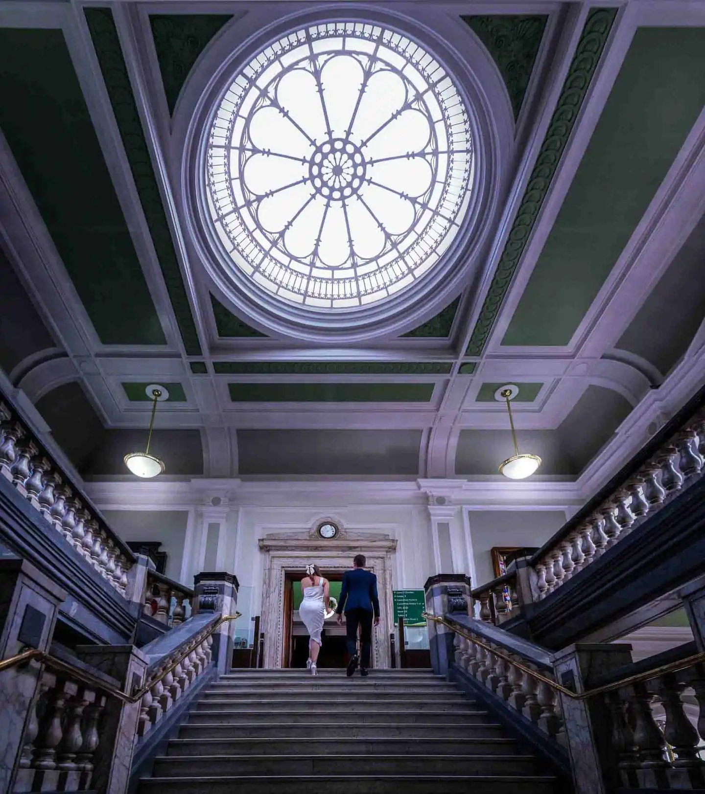 A bride and groom walk up a grand marble staircase toward a circular stained glass skylight.