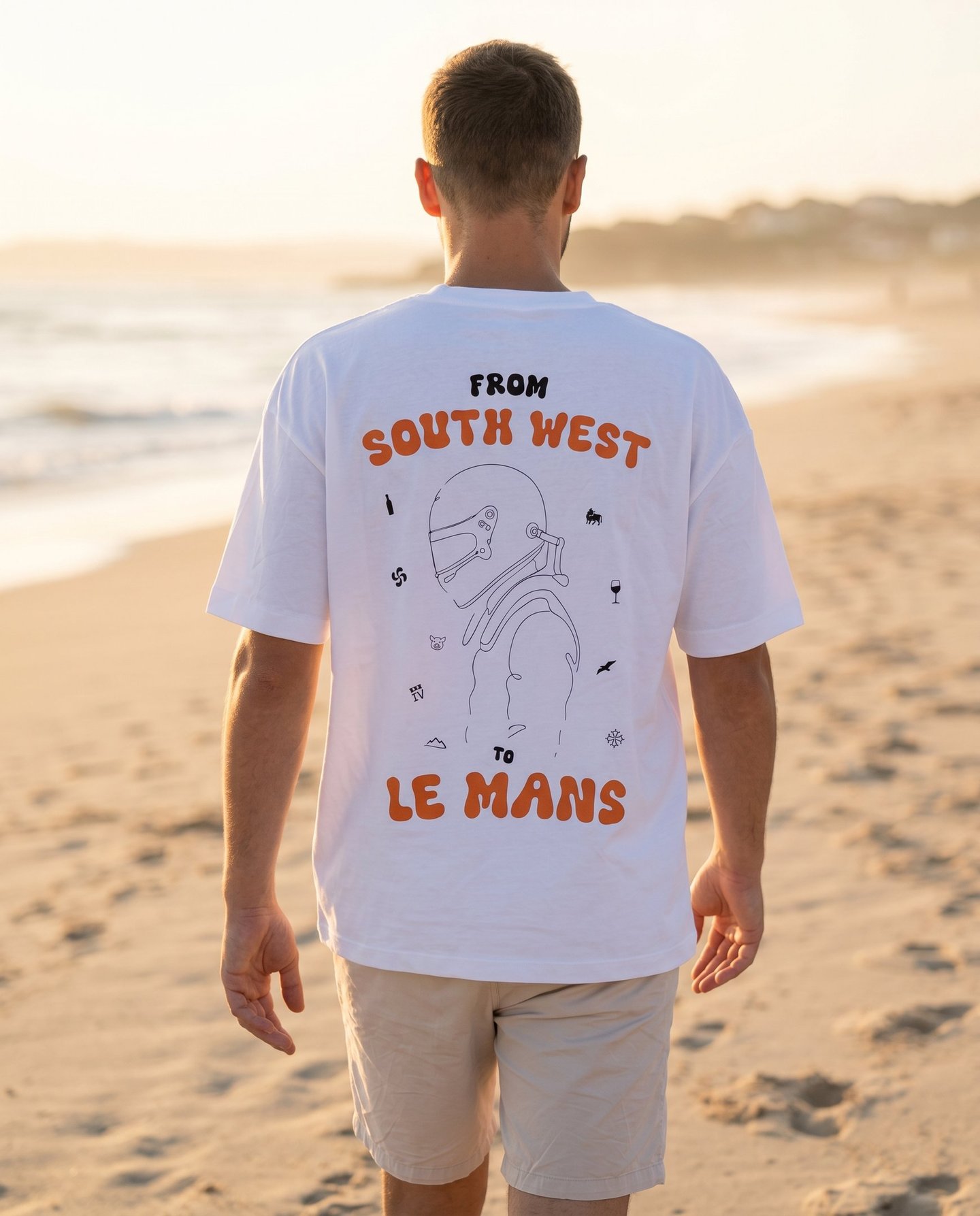 A man wearing a white graphic t-shirt with orange text and a racing driver illustration on a beach.