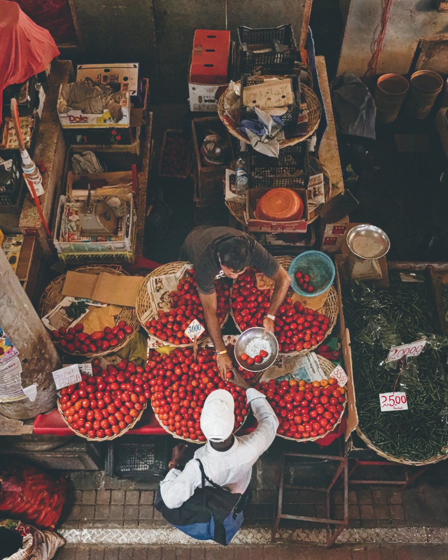 Overhead view of two men interacting at a market stall with baskets full of tomatoes, surrounded by various produce