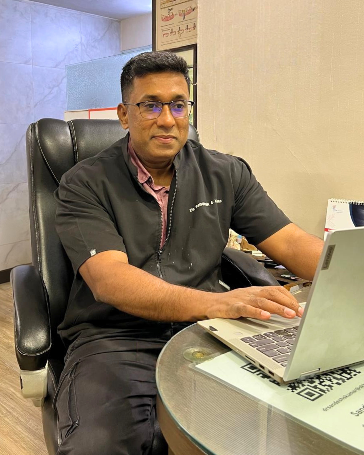 Male healthcare professional in black scrubs working on a laptop at his office desk.