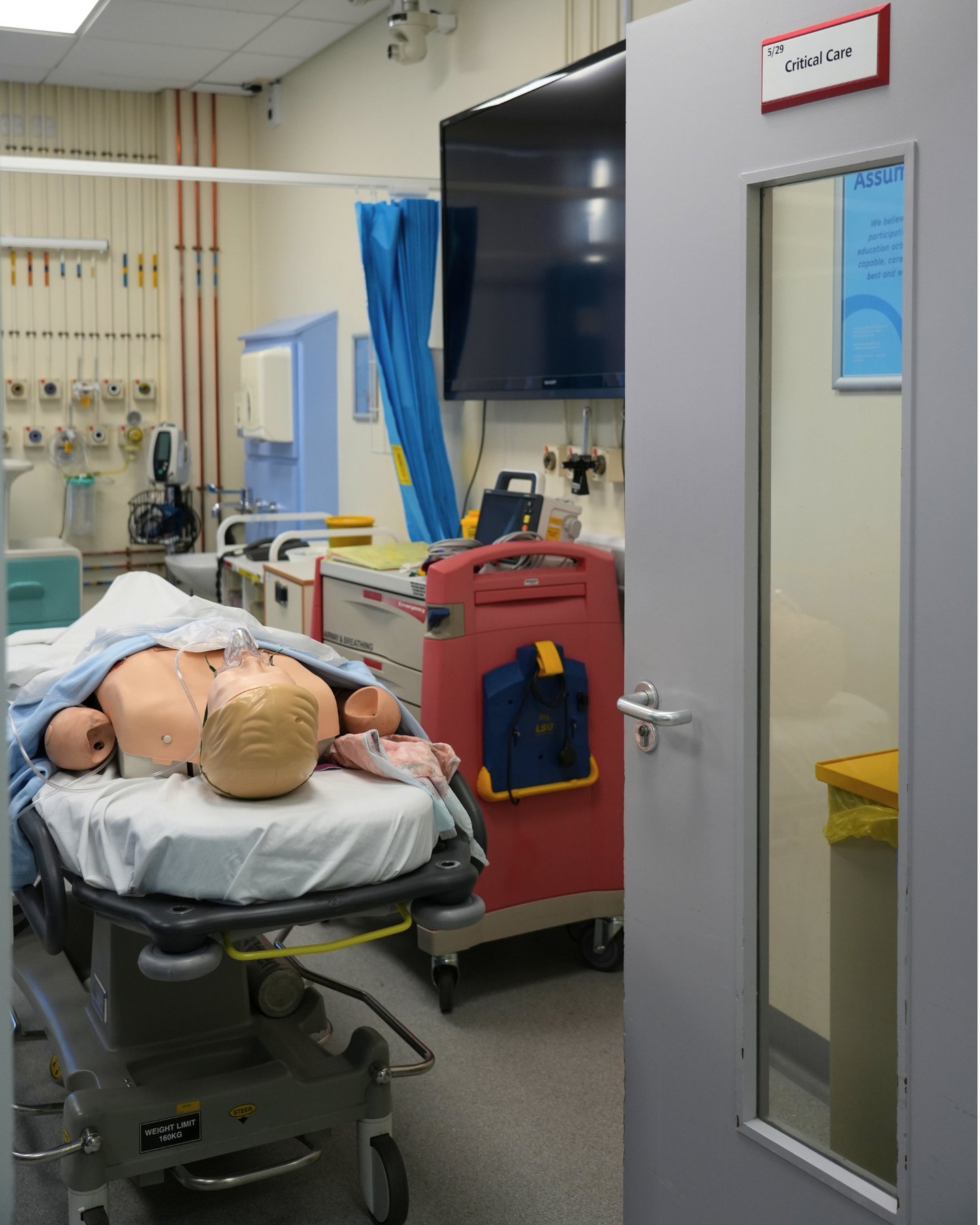 Medical simulation mannequin on a stretcher in a critical care training room with hospital equipment.