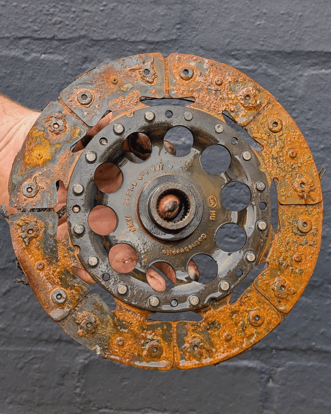 Hand holding a heavily rusted and worn automotive clutch disc against a dark wall background.