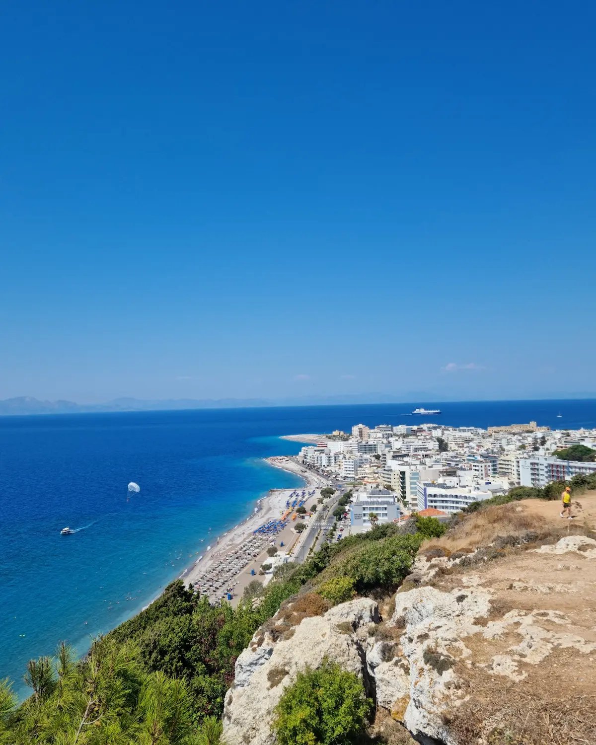 Aerial view of Rhodes City beach with white buildings, blue Aegean Sea, and parasailing on a sunny day.