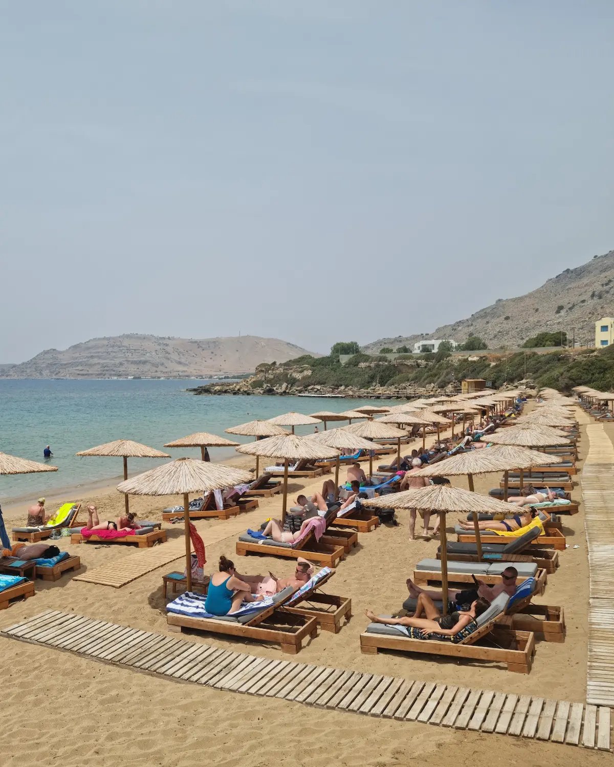 Tourists relaxing on wooden sunbeds under straw umbrellas at a sunny Mediterranean beach resort.