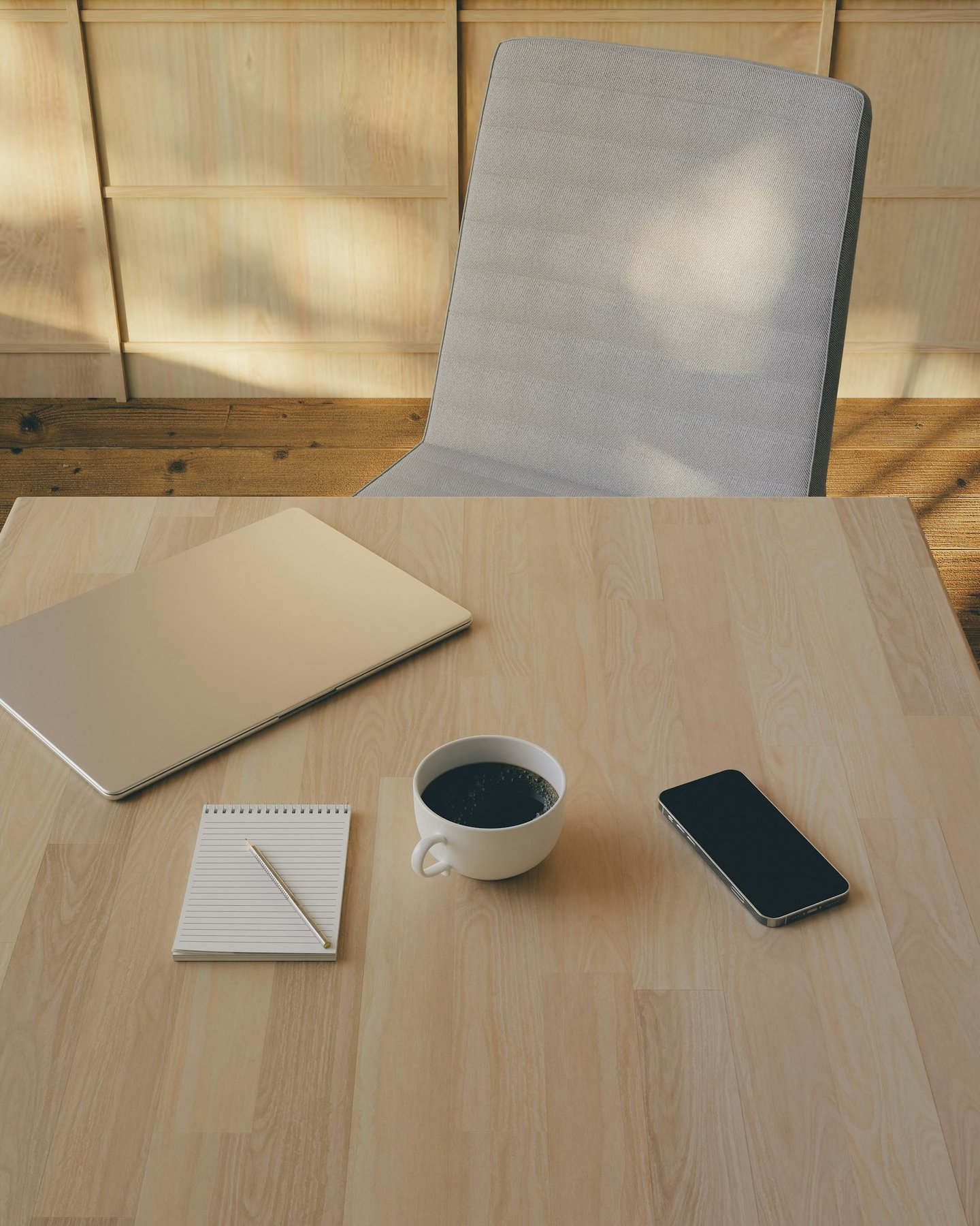 A modern workspace with a white chair in front of a desk iwith a closed laptop, notepad and pen, coffe cup, and smartphone.