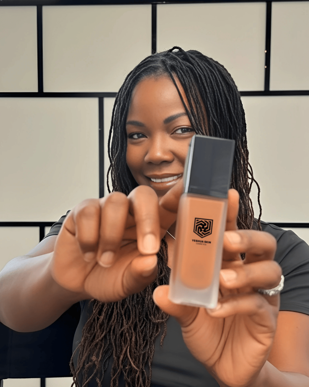 A woman holding a bottle of Yeshua Skin liquid foundation for dark skin tones.