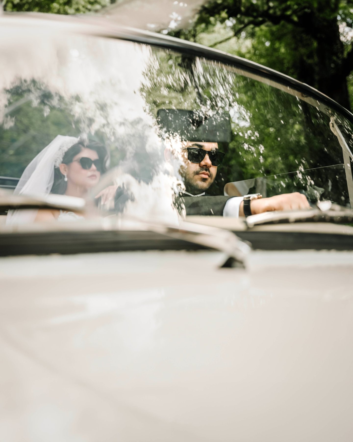 Bride and groom wearing sunglasses driving a classic car, captured by Fred Art Studio.