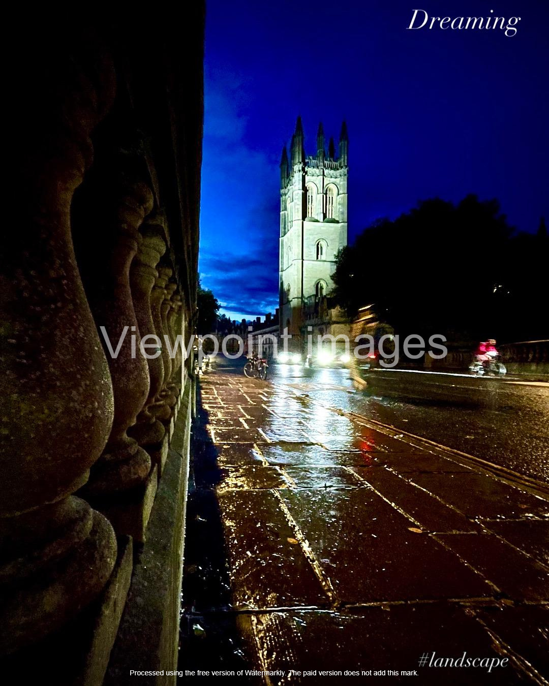 Magdalen College Tower at night viewed from a rainy Oxford street with reflections on wet pavement.