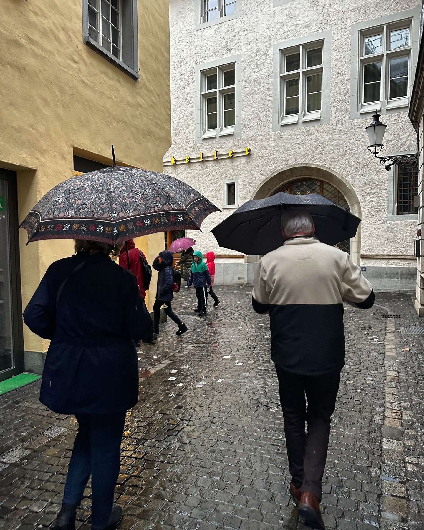 Rainy day in Switzerland, an older couple walk down the wet cobblestone roads together by LV