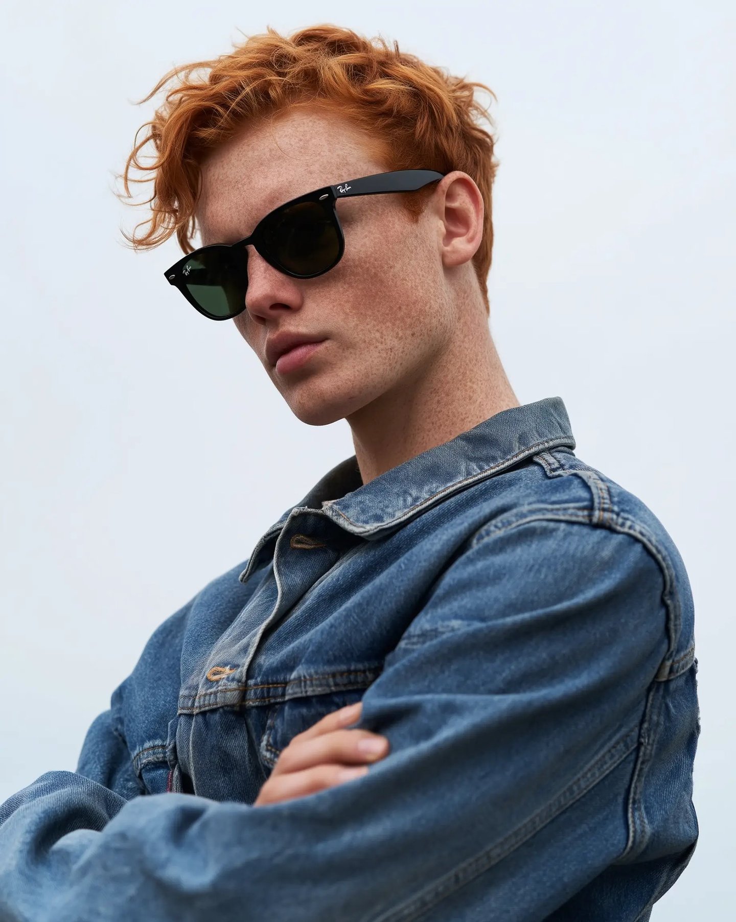 Young man with curly red hair and freckles wearing black Ray-Ban sunglasses and a blue denim jacket.
