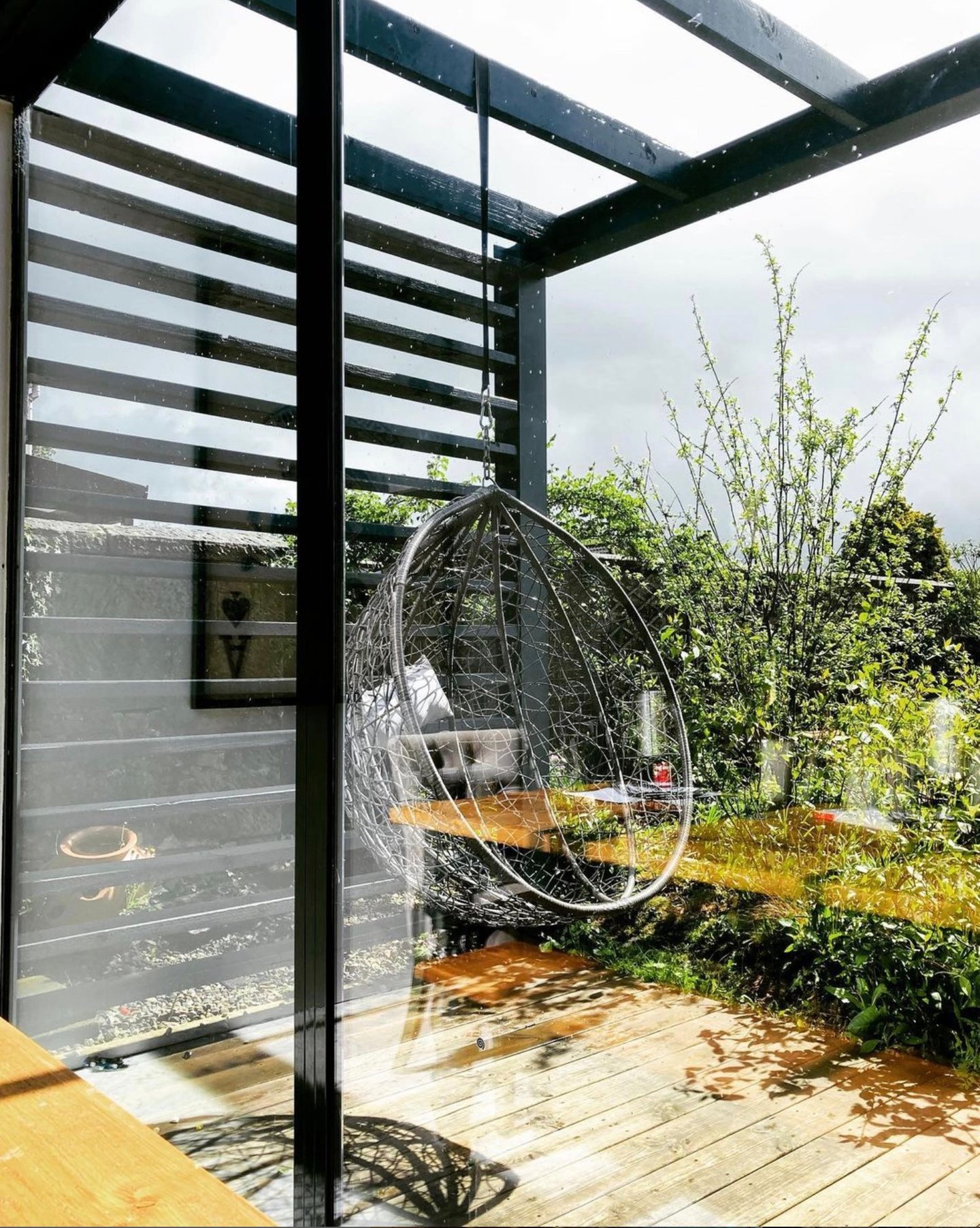 Modern hanging egg chair on a wooden deck under a black pergola in a lush garden patio.