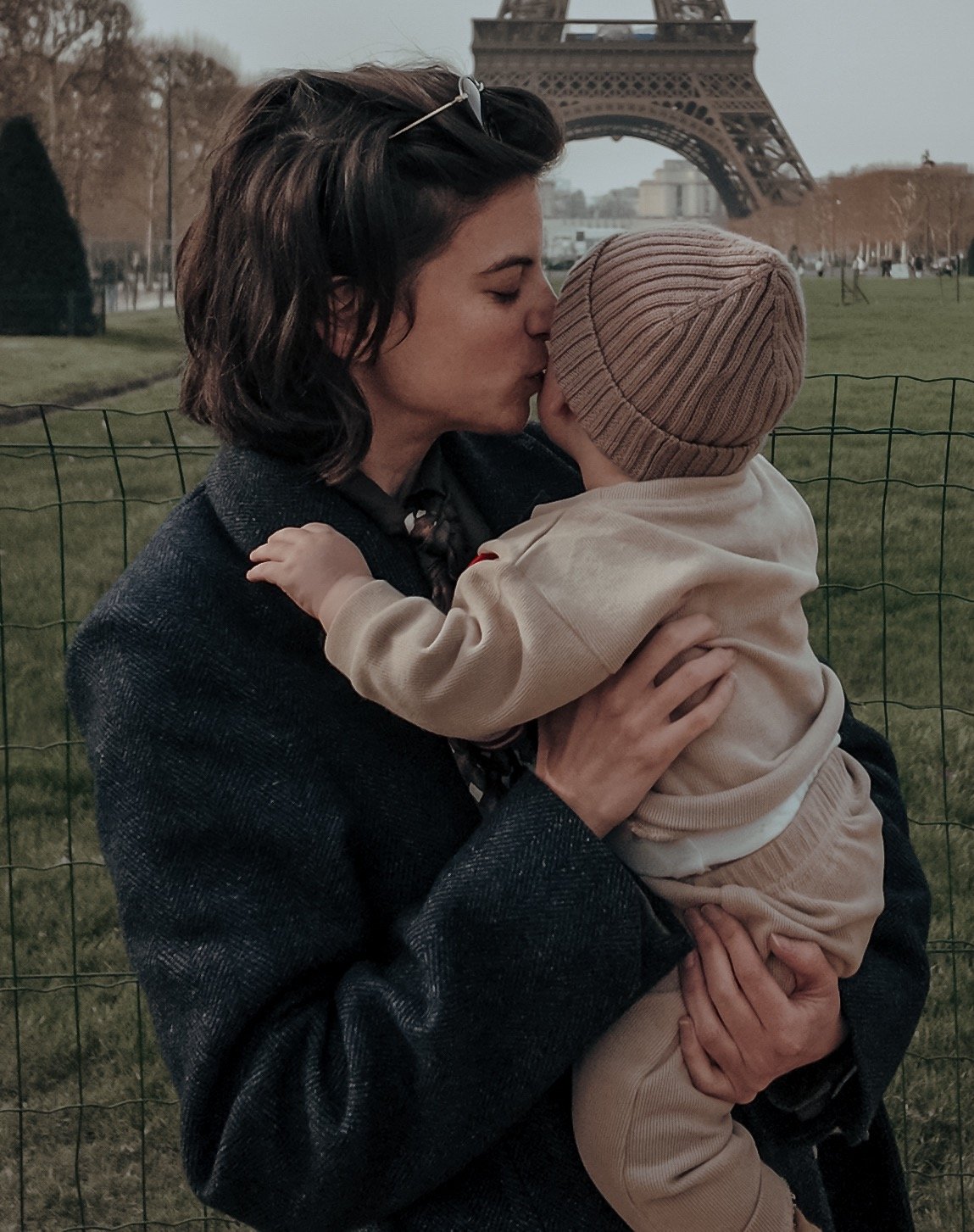 Un parent portant un manteau tient un jeune enfant en tenue beige près de la Tour Eiffel, montrant u