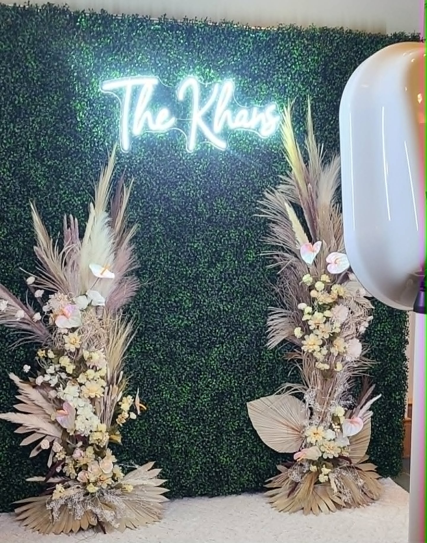 A wedding photo booth backdrop with a neon sign, green boxwood wall, and dried floral arrangements.