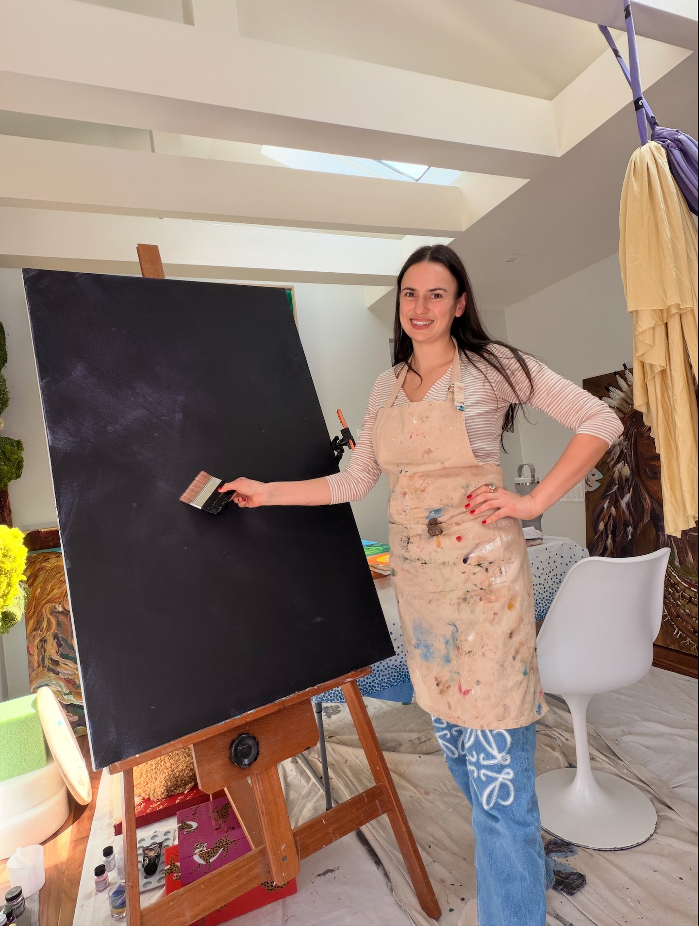 artist in a paint-stained apron holds a brush against a large black canvas in her studio.