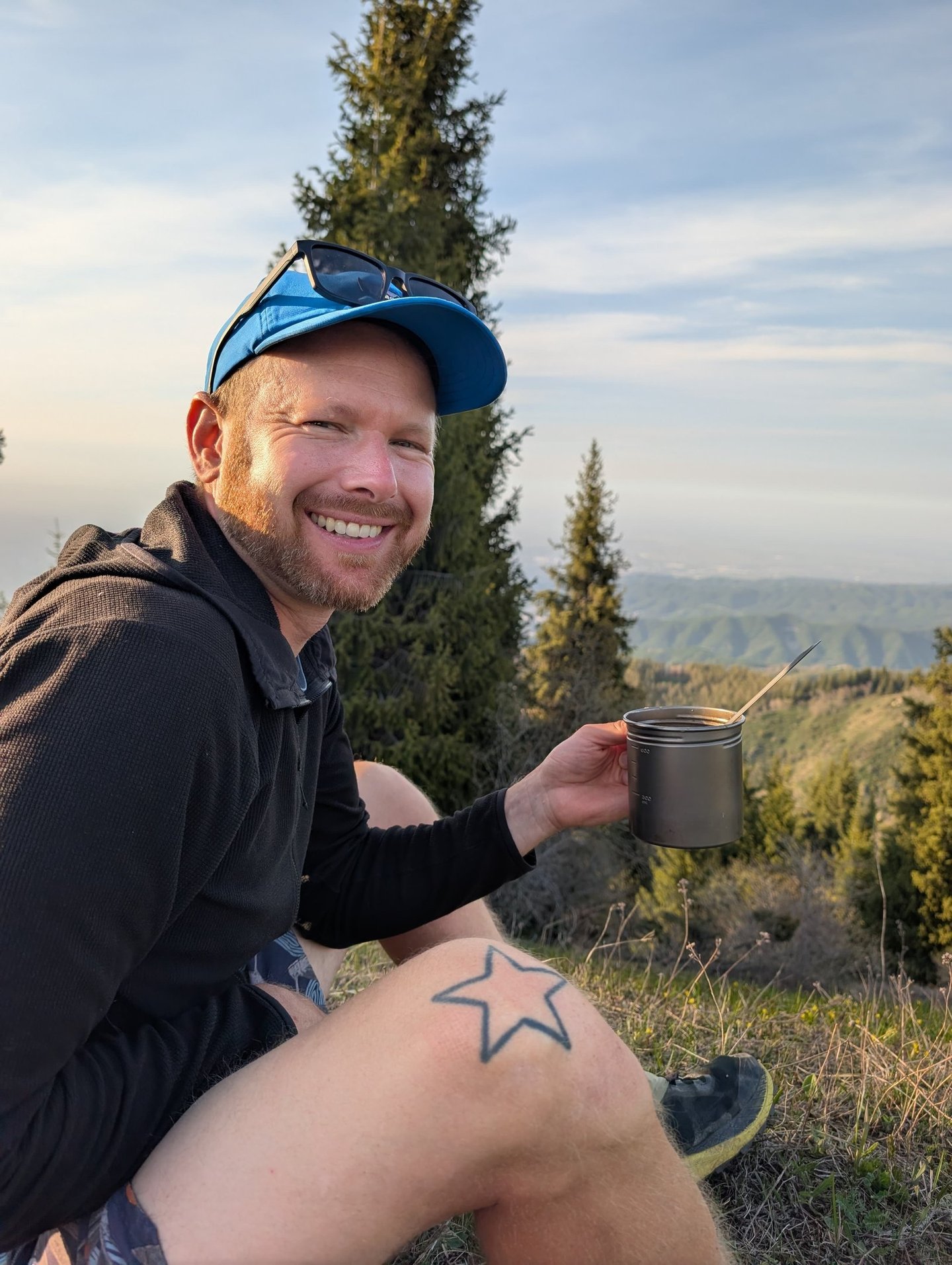 A smiling male hiker holding a Vargo BOT 700 mug while sitting on a scenic mountain ridge at sunset.