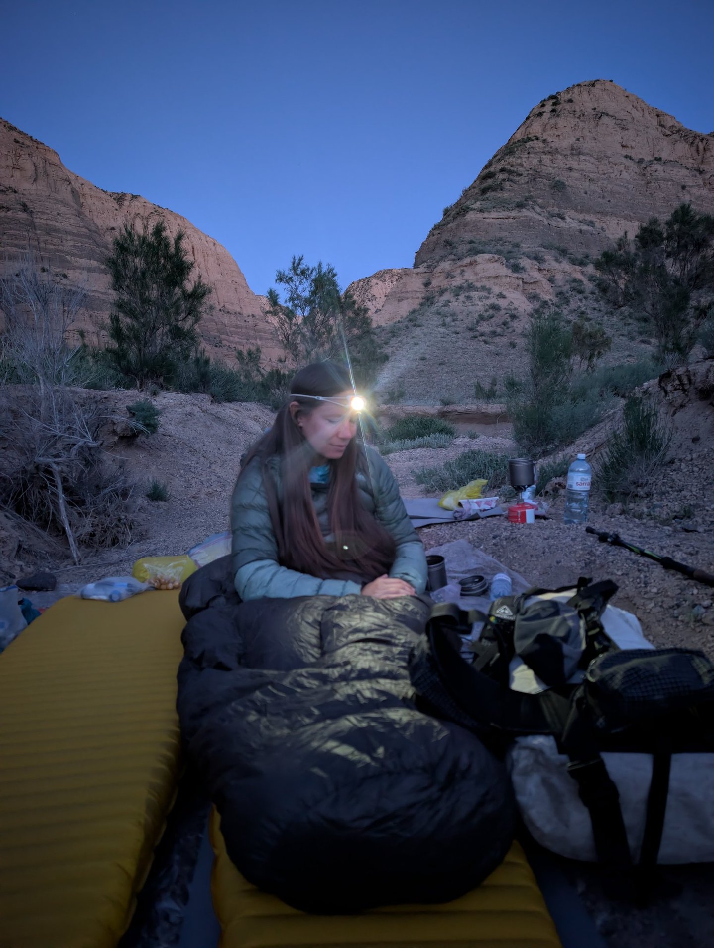 Bugs in her sleeping bag using her Petzl Bindi headlamp as dusk in a desert canyon campsite.