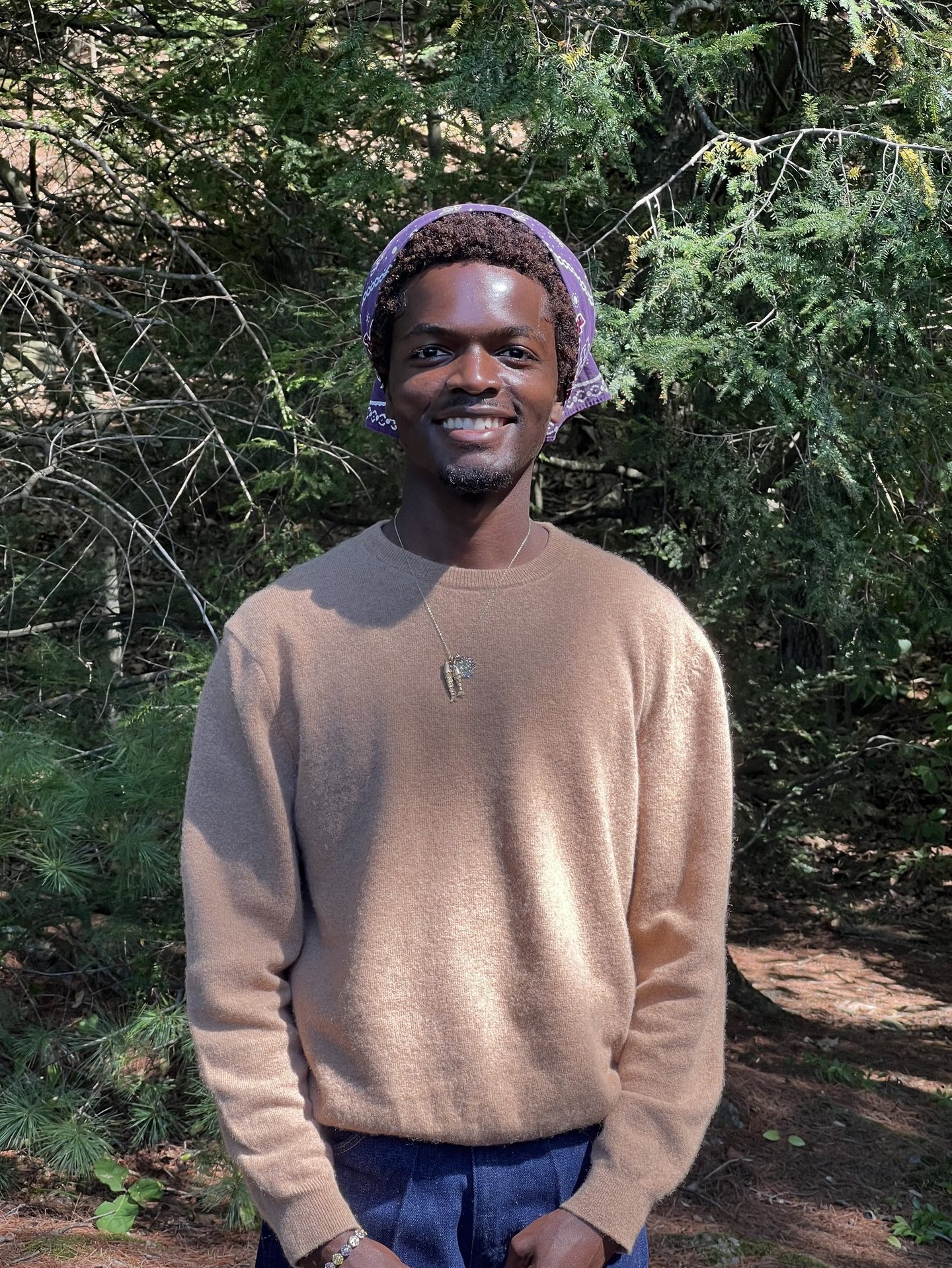 A smiling man wearing a beige sweater and purple bandana standing in a lush green forest.
