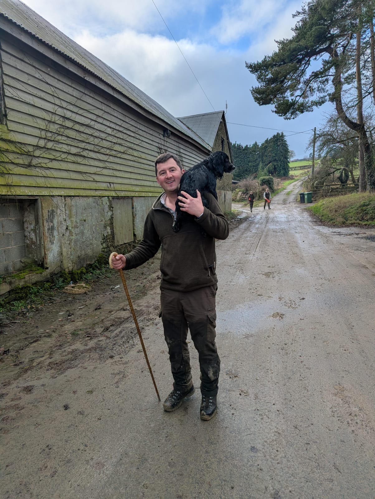 A smiling man in country attire carries a black spaniel dog on his shoulder along a rural farm track.