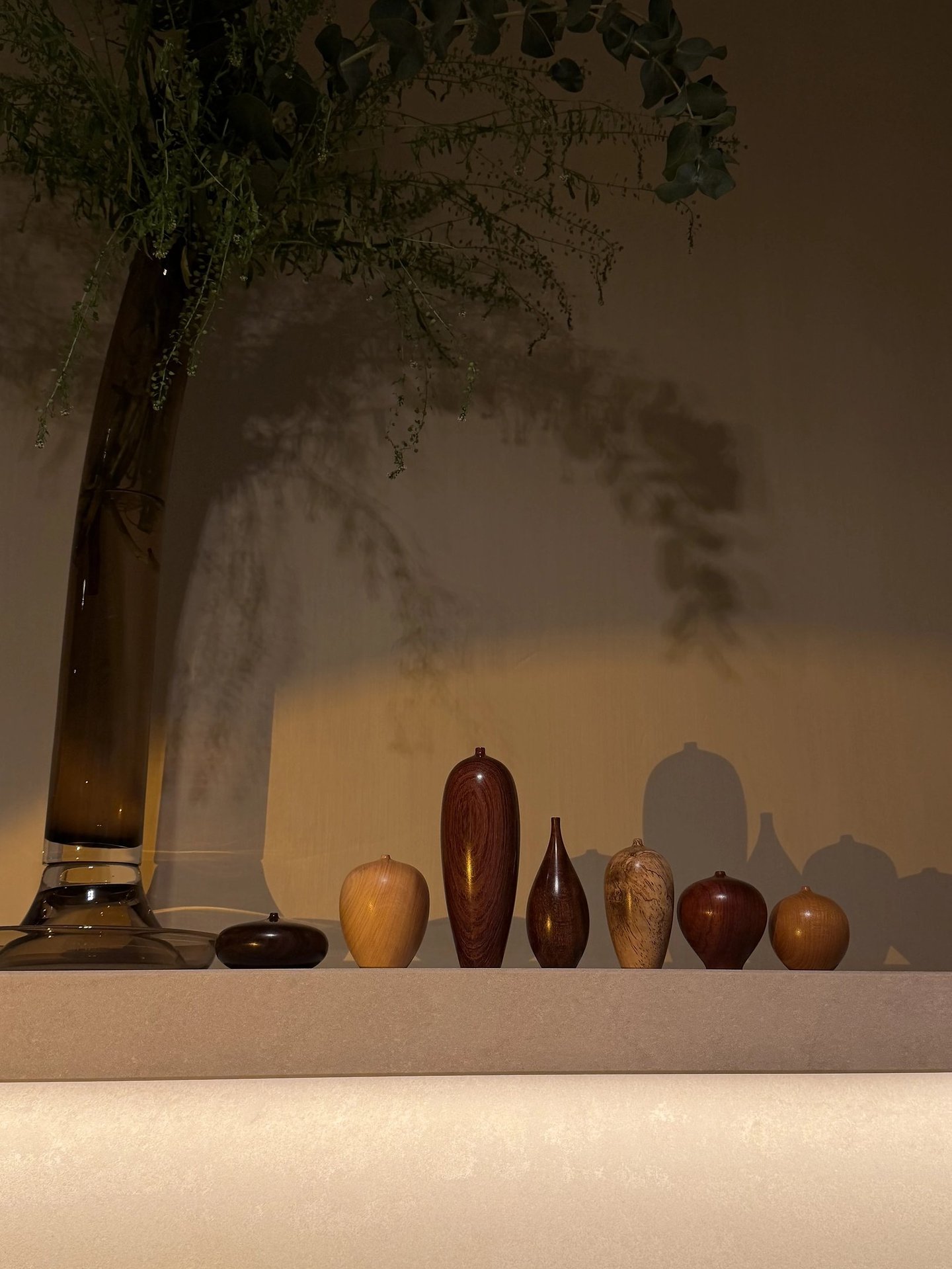 Hand-turned wooden vessels and a glass vase with eucalyptus branches displayed on a shelf with shadows.