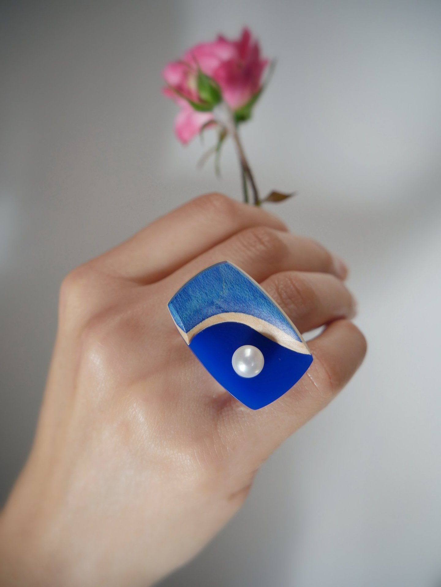 Hand wearing a blue statement wooden ring with a white pearl accent while holding a pink rose.